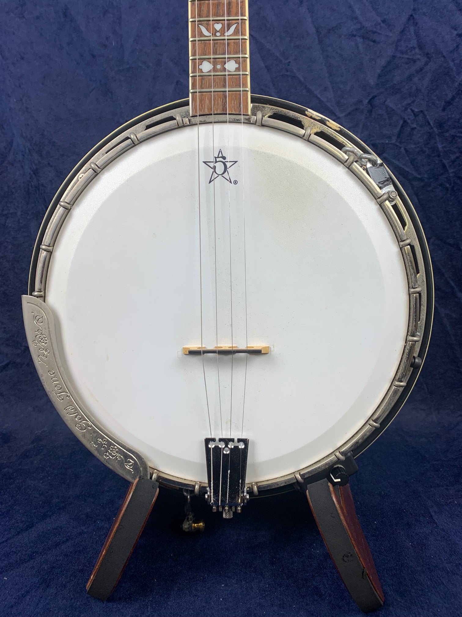 Pre-owned vintage banjo with a white drumhead, metal rim, wooden neck, and decorative inlays; sits upright on a stand against a dark background.