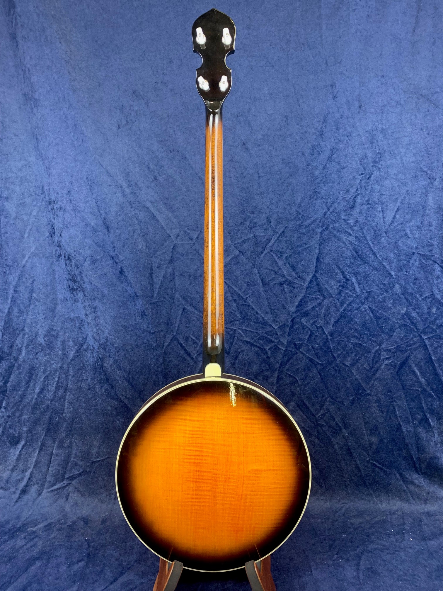 Vintage banjo shown from the back, featuring a sunburst finish on the round body and a straight wooden neck against a blue background.