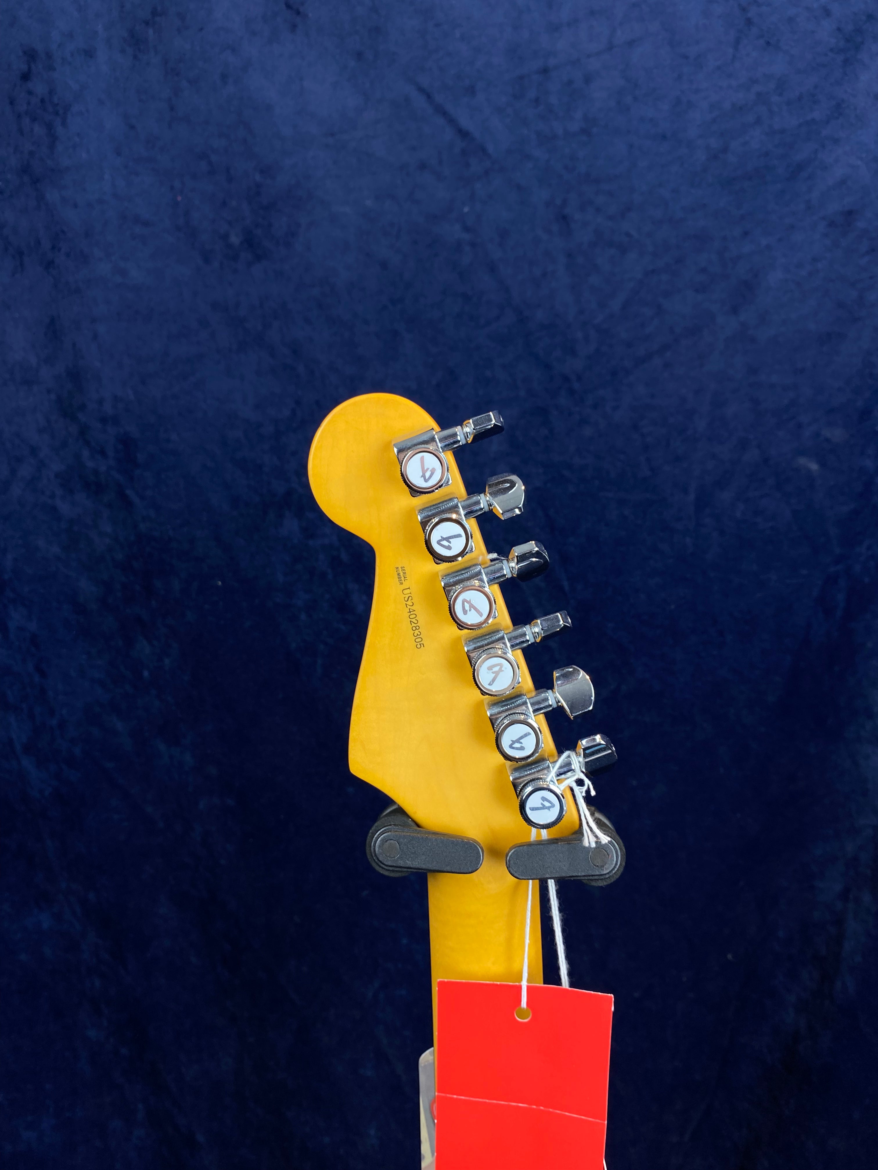 Back of an electric guitar headstock showing six chrome tuning pegs, serial number, yellow wood finish, and red hang tag against a dark blue background.