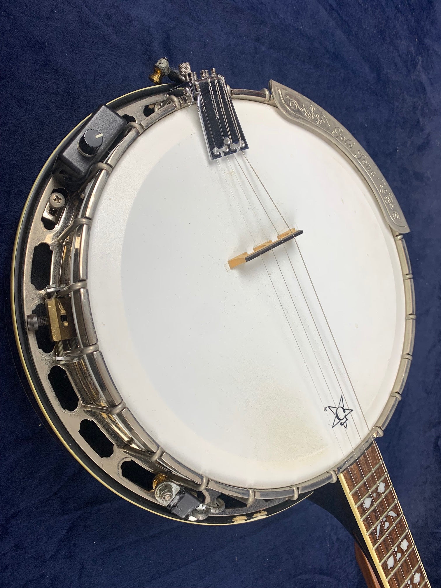 Vintage five-string banjo with engraved metal armrest, wooden neck, and white drum head; visible wear, electronic pickup attached.