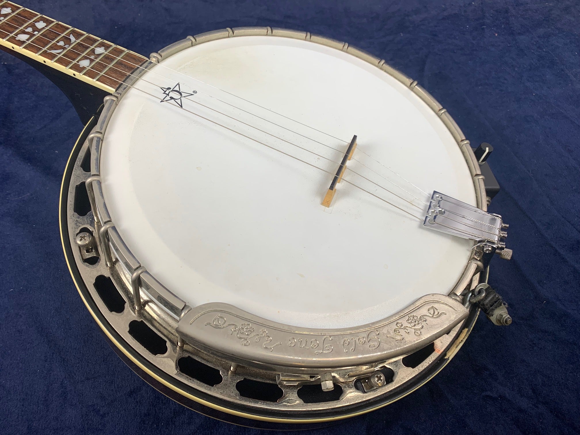 Vintage pre-owned banjo with a white drumhead, metal rim, and engraved armrest, shown on a dark blue cloth background.