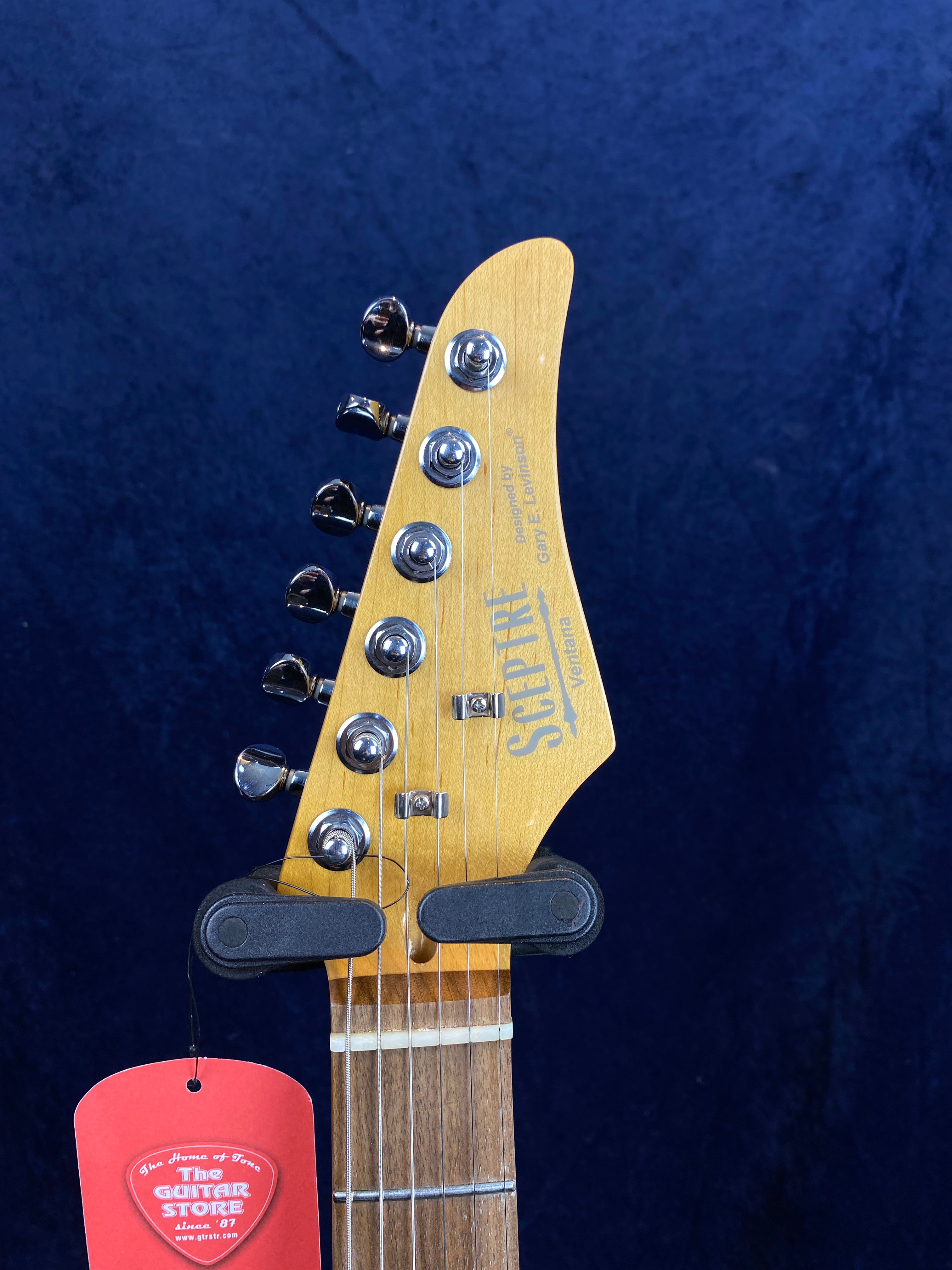 Sceptre electric guitar headstock with six chrome tuning pegs, light wood finish, and a red tag labeled "The Guitar Store" hanging from a string.