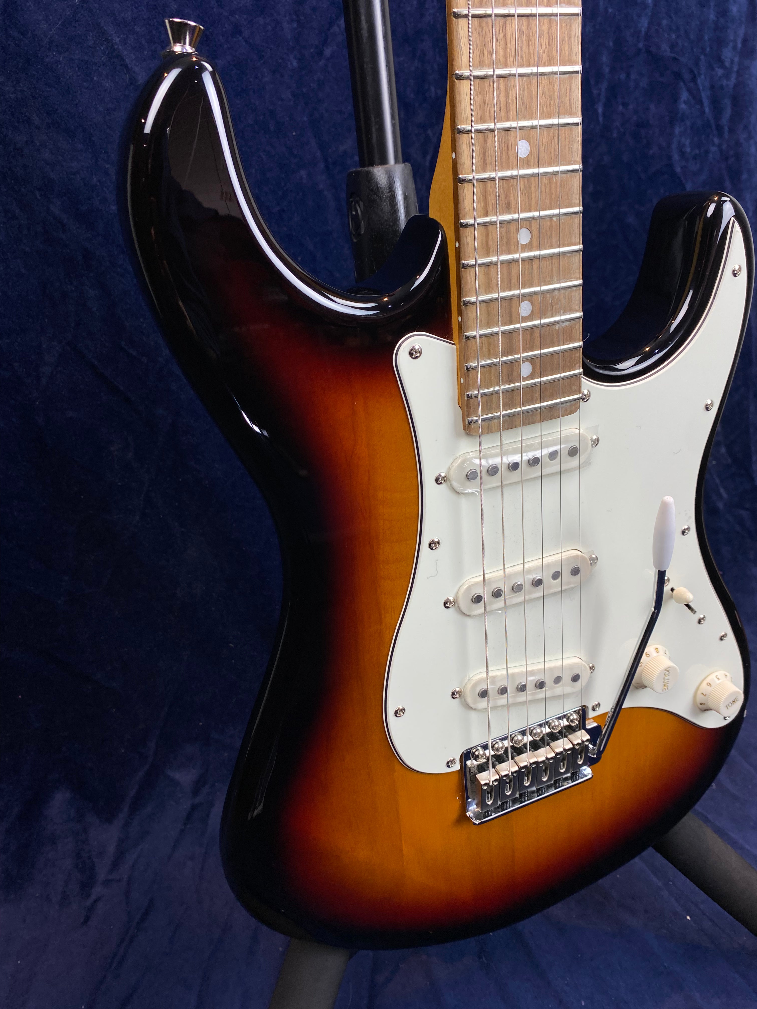 Sunburst electric guitar with a white pickguard, three single-coil pickups, control knobs, and a tremolo bar, shown against a dark background.