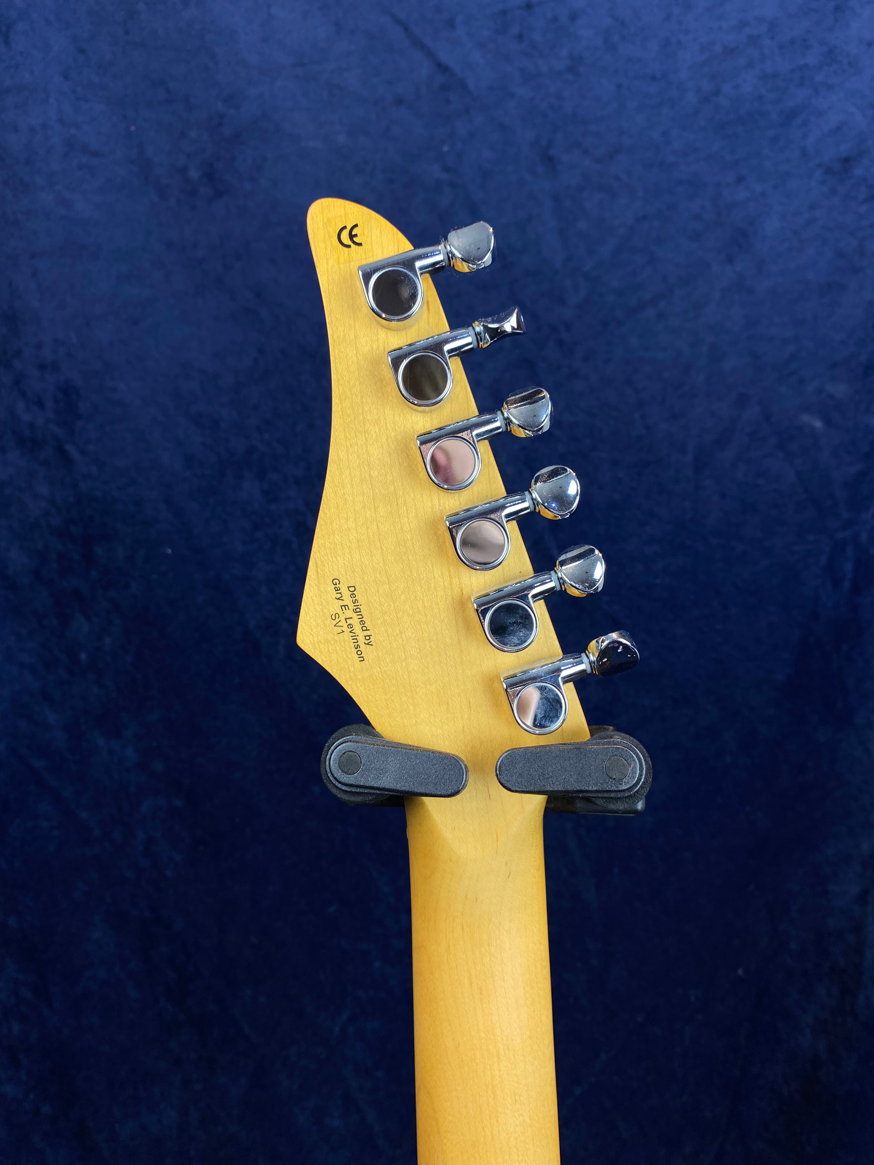 Six chrome tuning pegs are lined up on the back of a light wood electric guitar headstock, with a guitar hanger holding the neck upright.