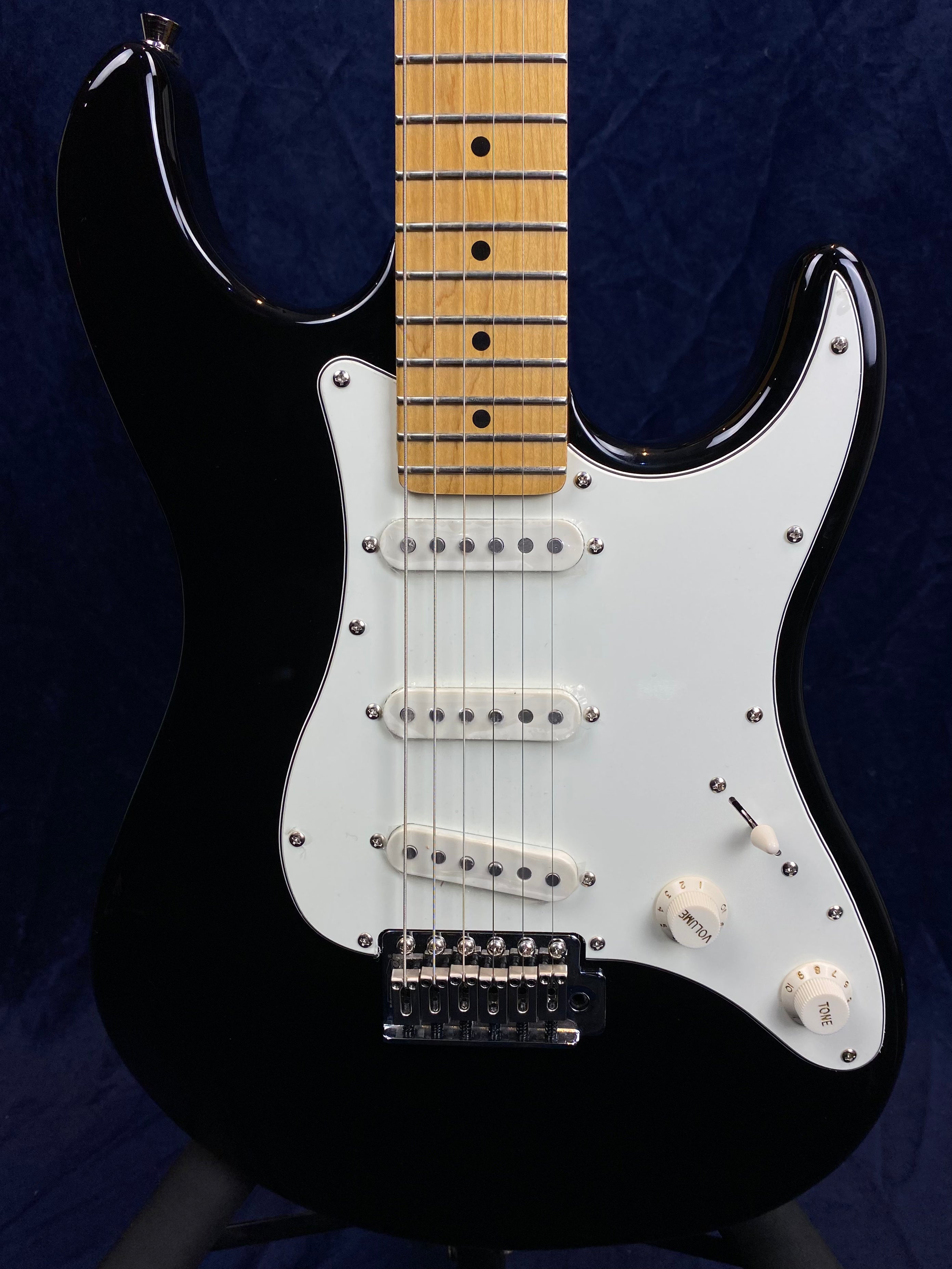 Black electric guitar with a white pickguard, three single-coil pickups, two control knobs, a switch, and a maple fretboard.