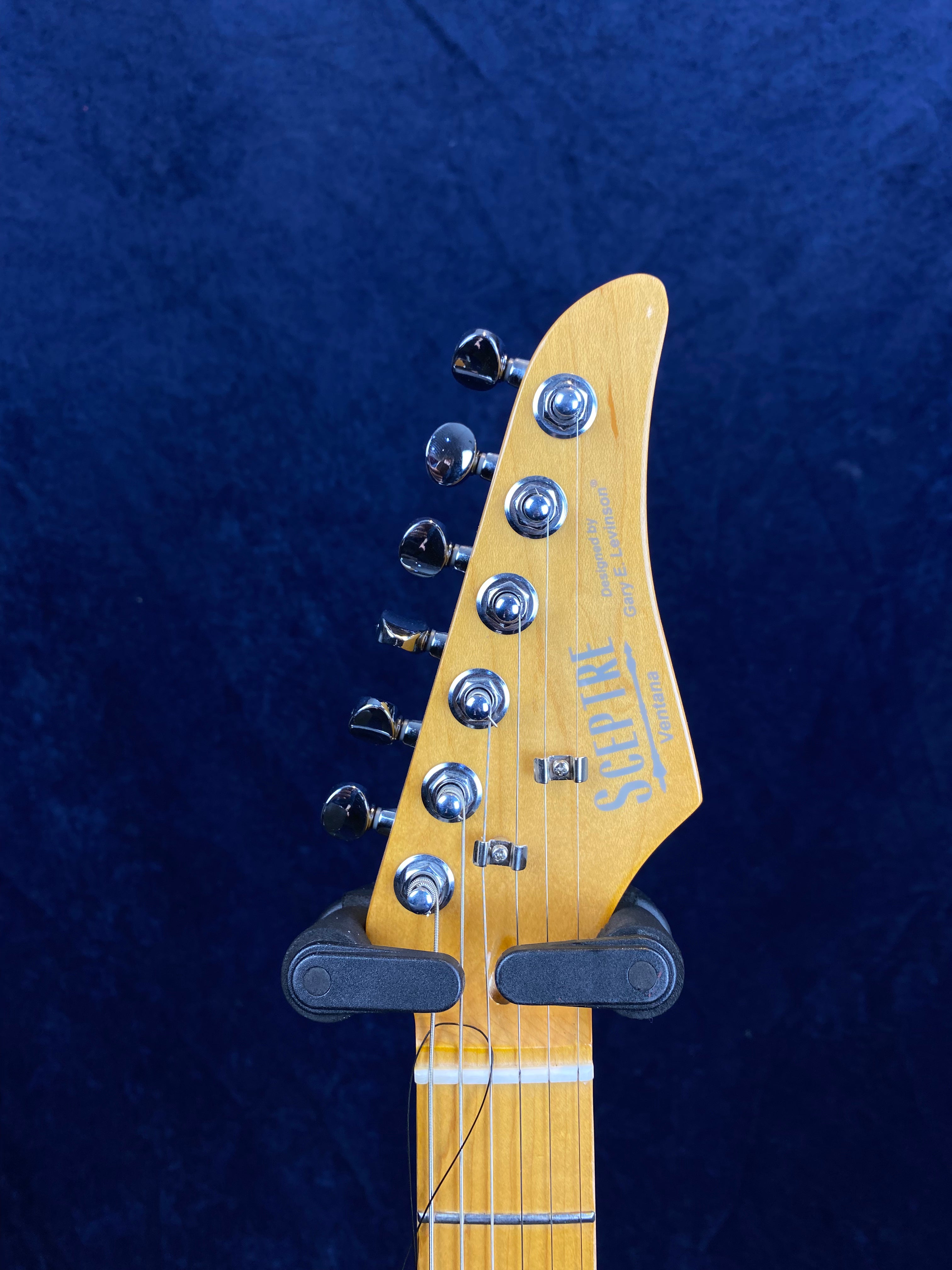 Light wood electric guitar headstock with six chrome tuning pegs and "Sceptre Ventura" logo, held in a black wall mount against a dark background.