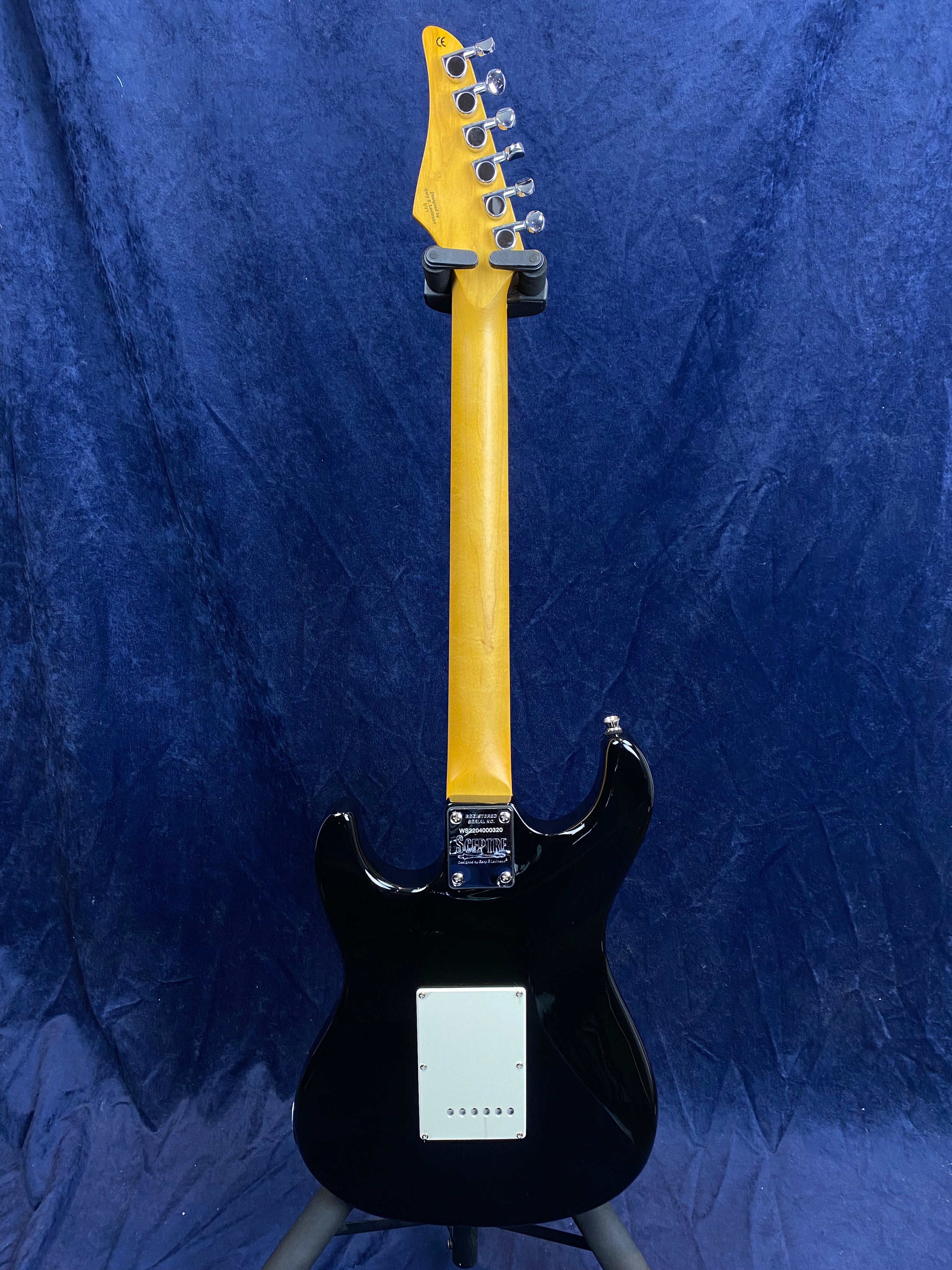 Black electric guitar shown from the back, with a natural wood neck, six tuning pegs, and a white plastic cover plate on the body.
