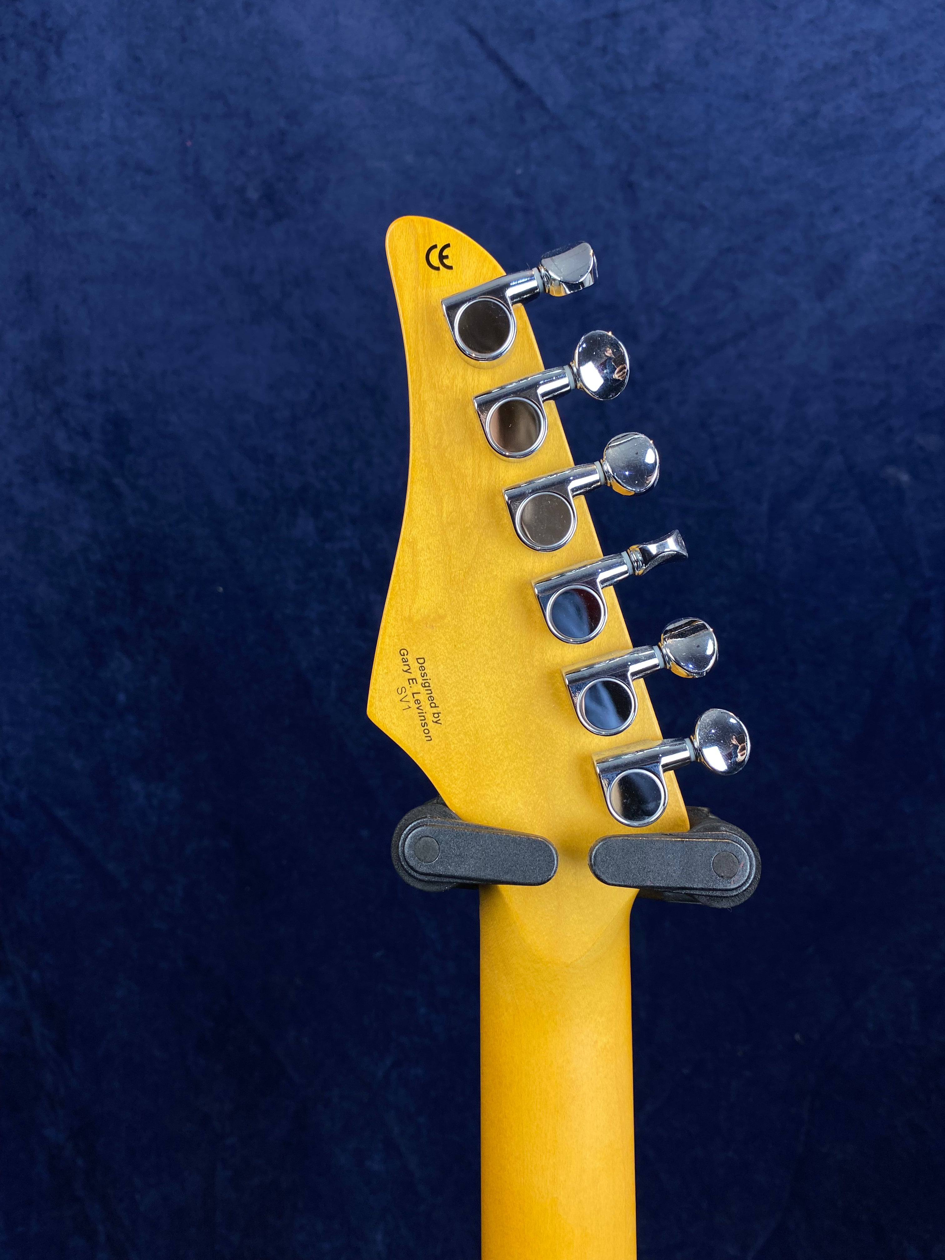 Six silver tuning pegs line the back of a light wood electric guitar headstock, shown mounted on a wall holder against a dark blue background.