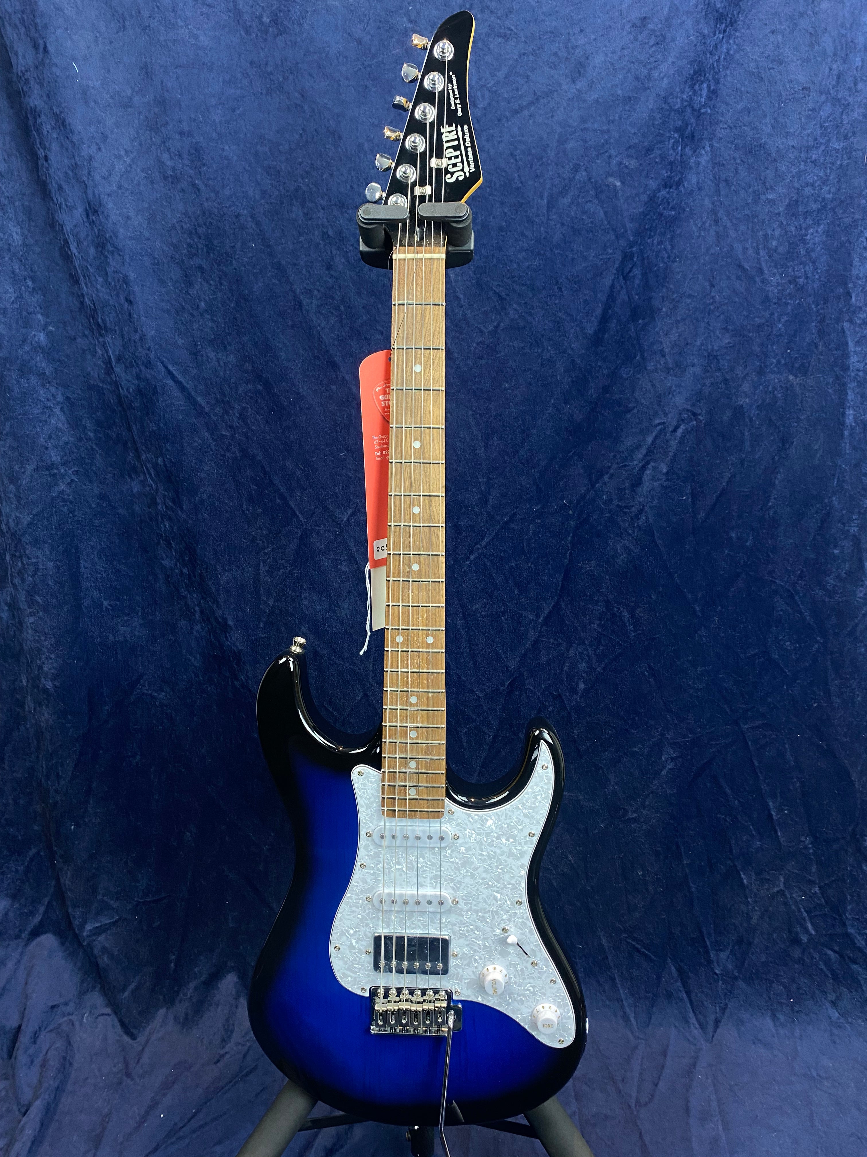 Electric guitar with a glossy blue and black sunburst body, white pearloid pickguard, maple neck, and six tuning pegs against a dark blue backdrop.