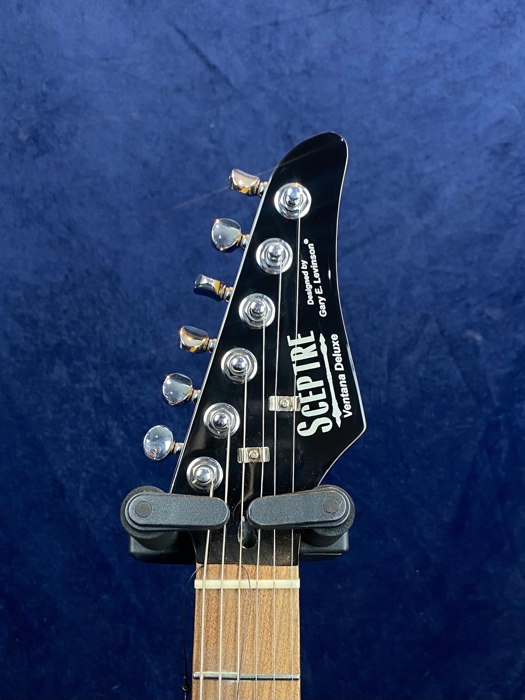 Close-up of an electric guitar headstock labeled Sceptre, showing six metal tuning pegs and part of a wooden neck against a blue background.