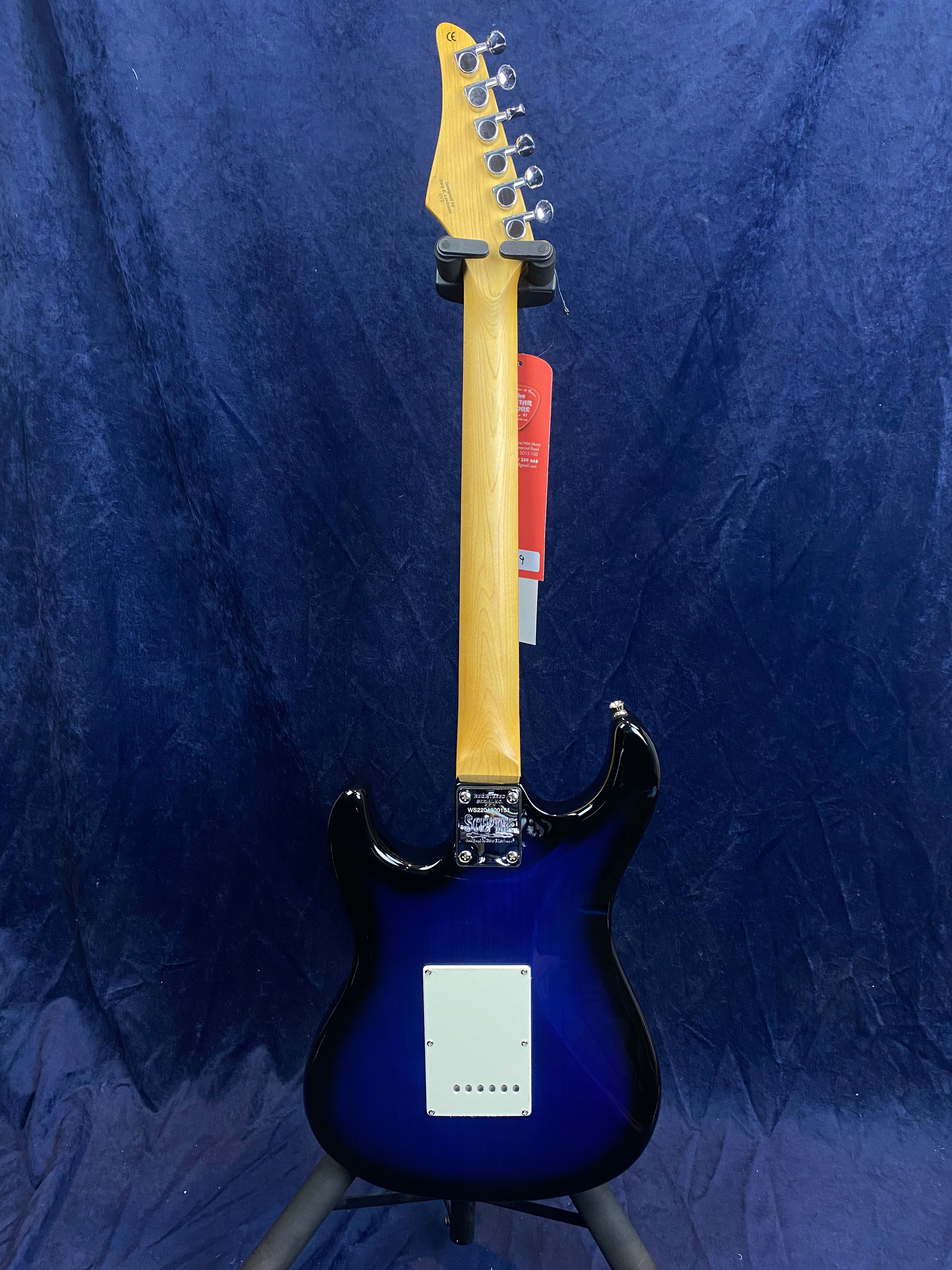 Blue electric guitar shown from the back, with a natural wood neck and white rectangular plate on the lower body, standing on a guitar stand.