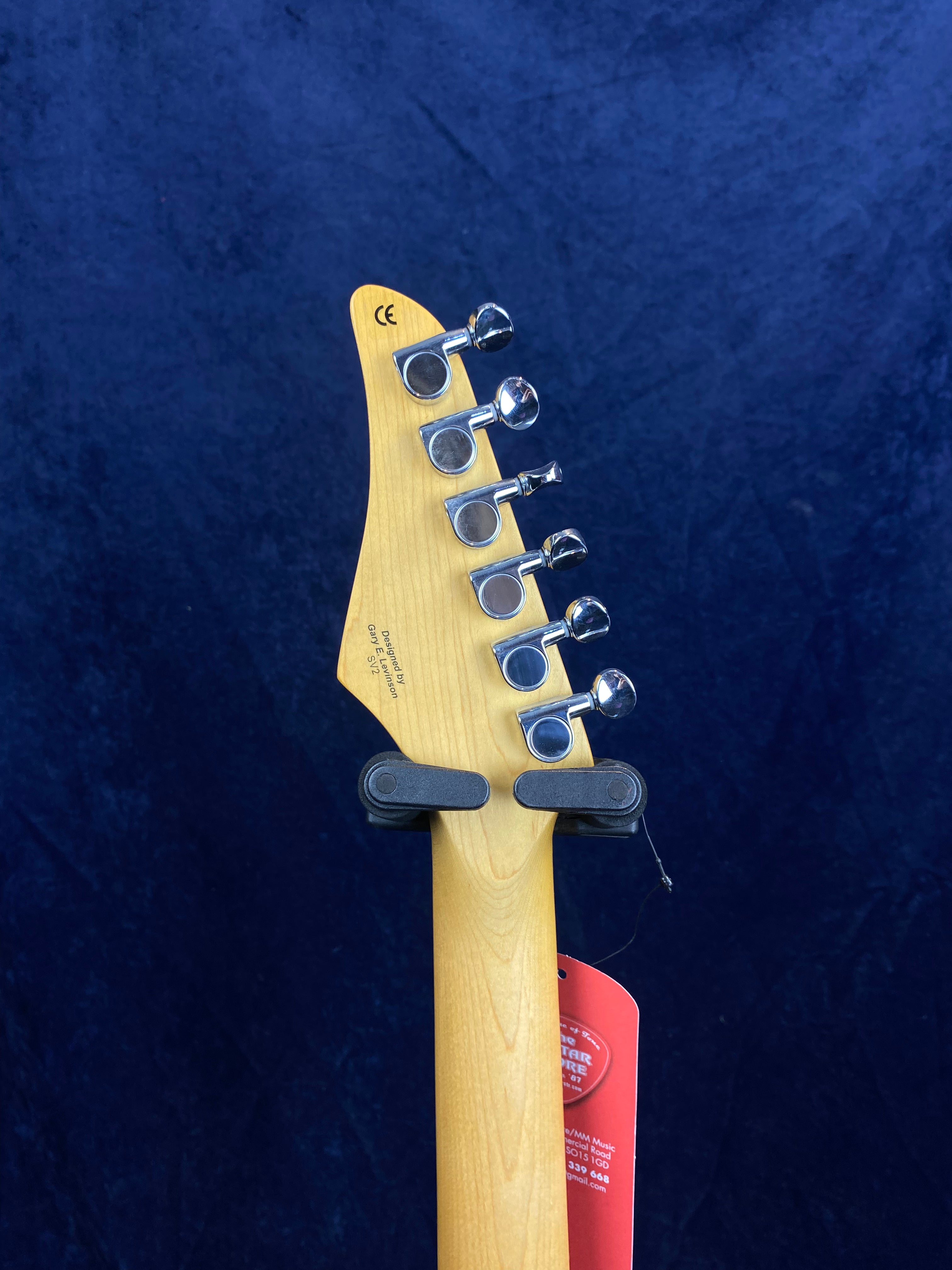 The back of an electric guitar headstock showing six metal tuning pegs, light wood finish, and a red retail tag hanging from the neck.