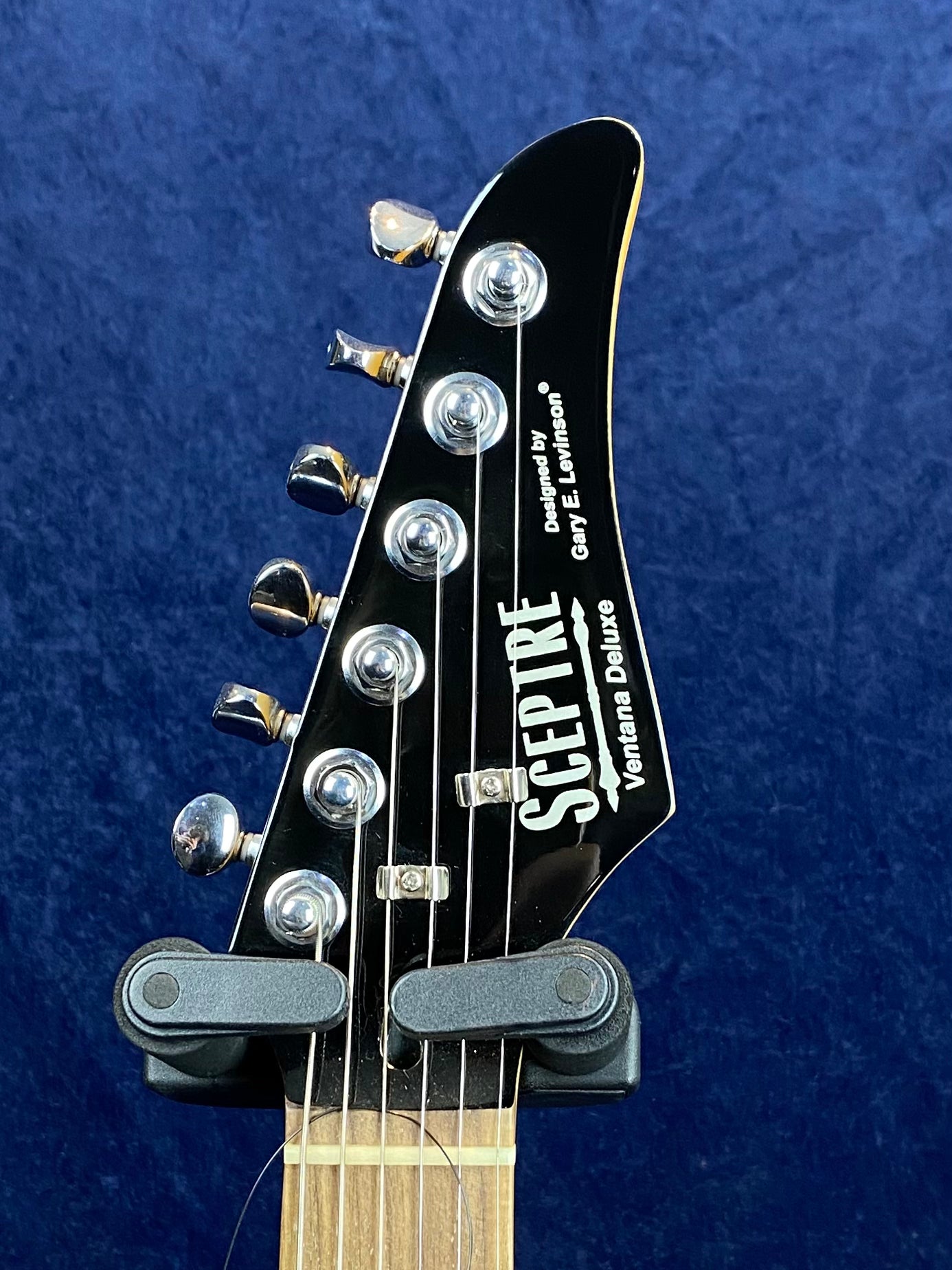 The headstock of a Sceptre Ventana Deluxe electric guitar with six tuning pegs and logo text, set against a dark blue background.