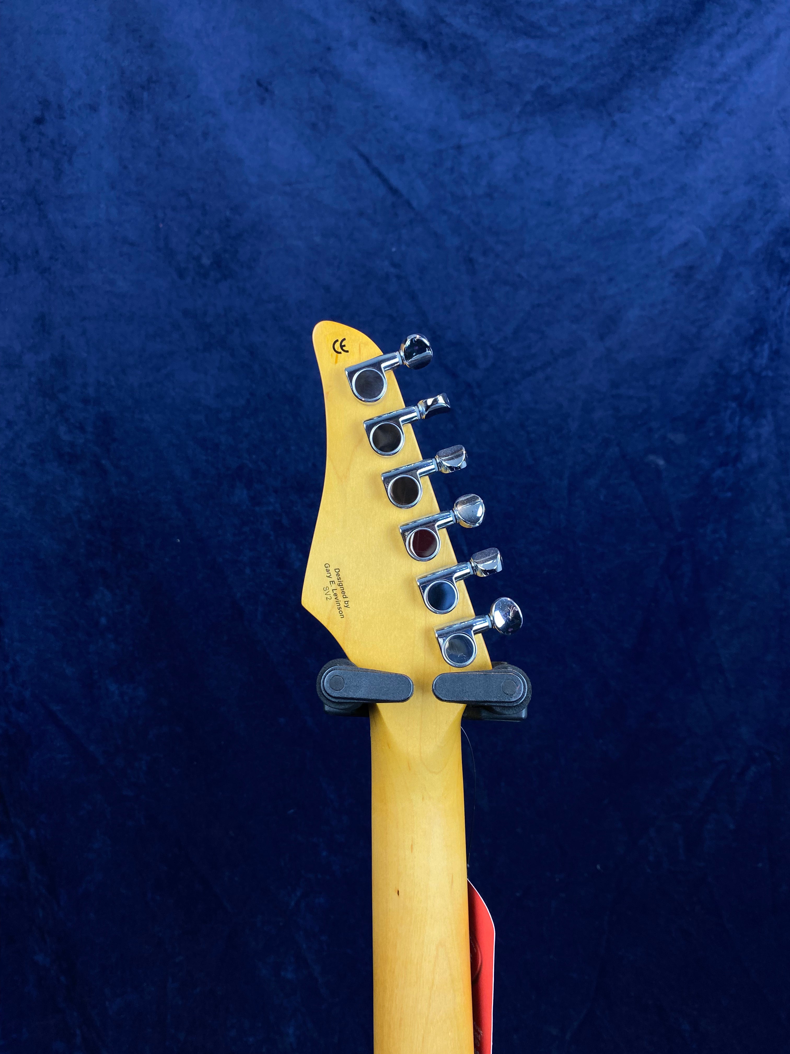 Six tuning pegs on the back of a light wood electric guitar headstock, held upright by a wall hanger, against a dark blue background.