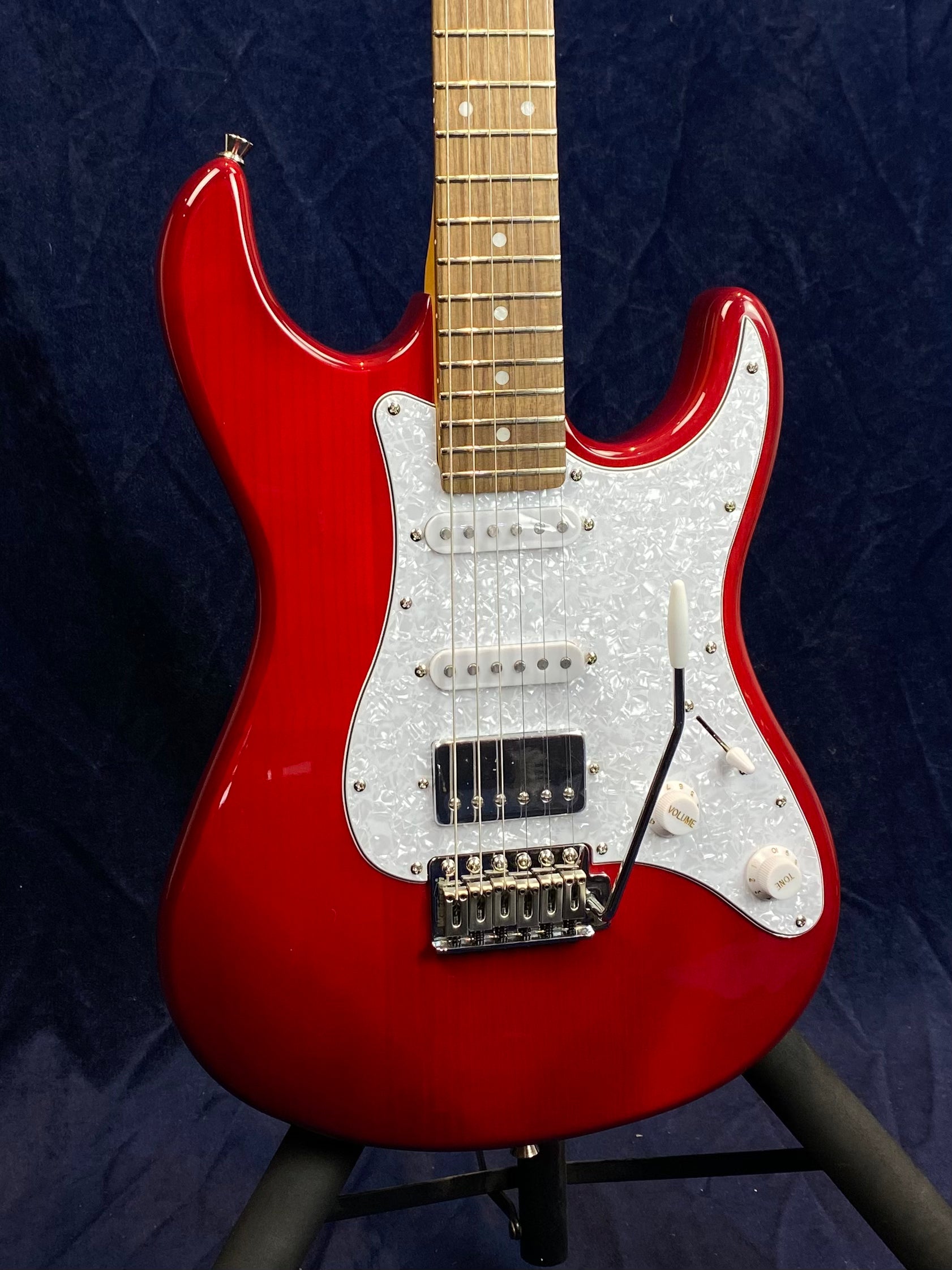 Red electric guitar with a white pearloid pickguard, three pickups, two control knobs, and a tremolo arm, displayed on a stand.