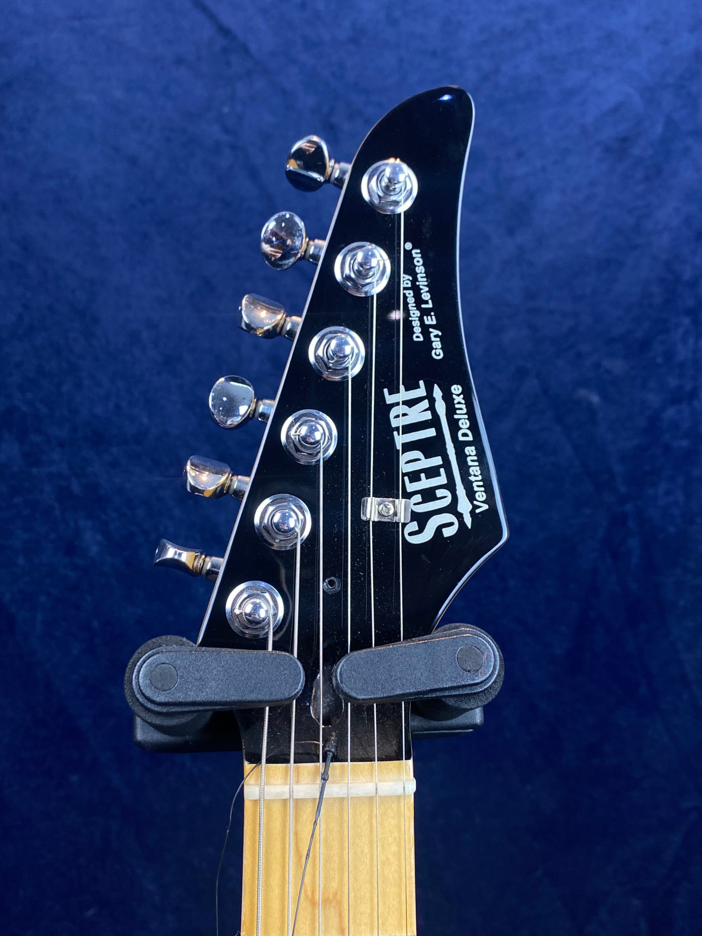 Close-up of a Sceptre Ventura Deluxe electric guitar headstock with six tuning pegs and a light wood neck against a dark blue background.