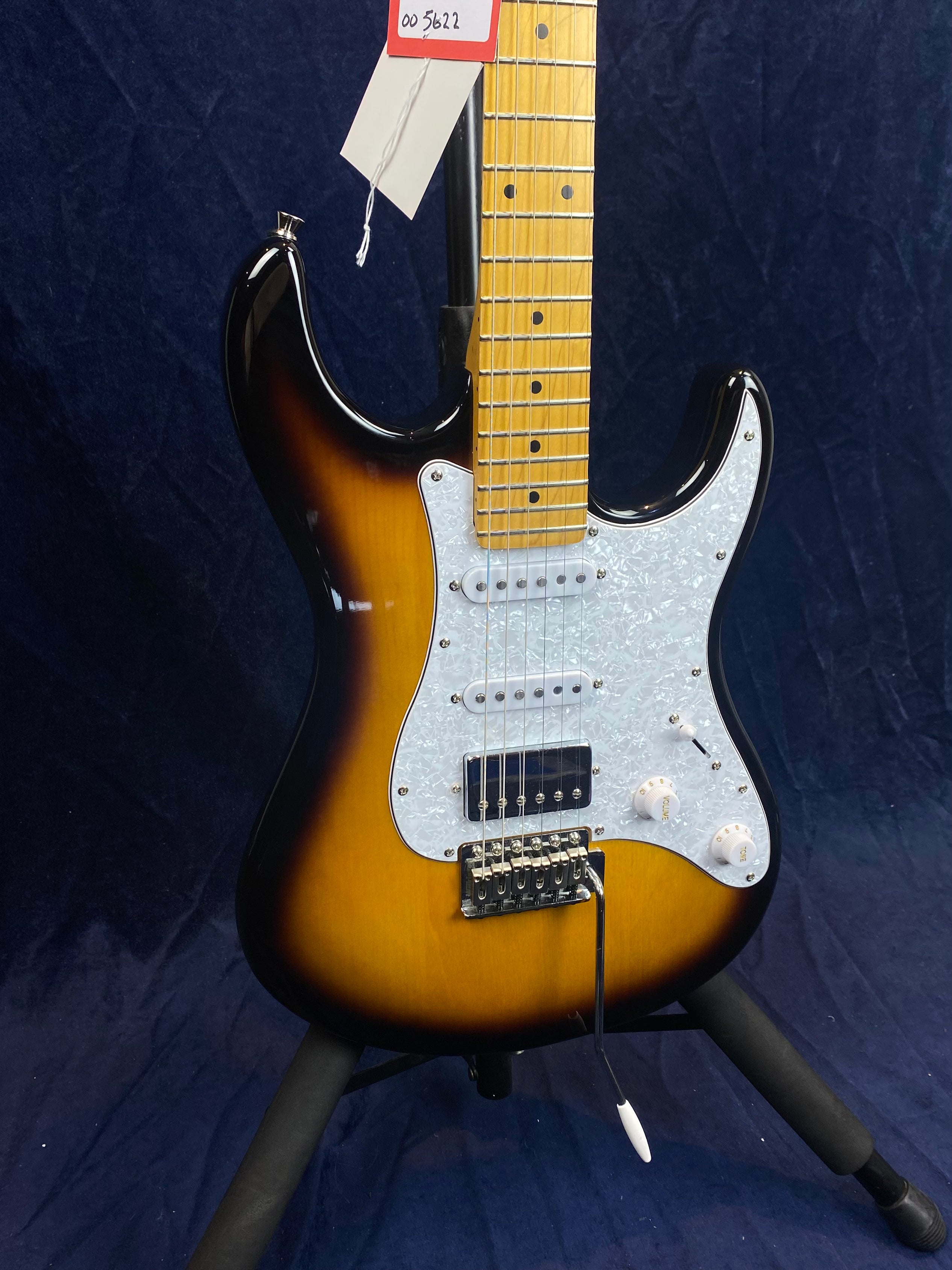 Sunburst electric guitar with a white pearloid pickguard, maple fretboard, three pickups, two control knobs, and a whammy bar, on a stand.