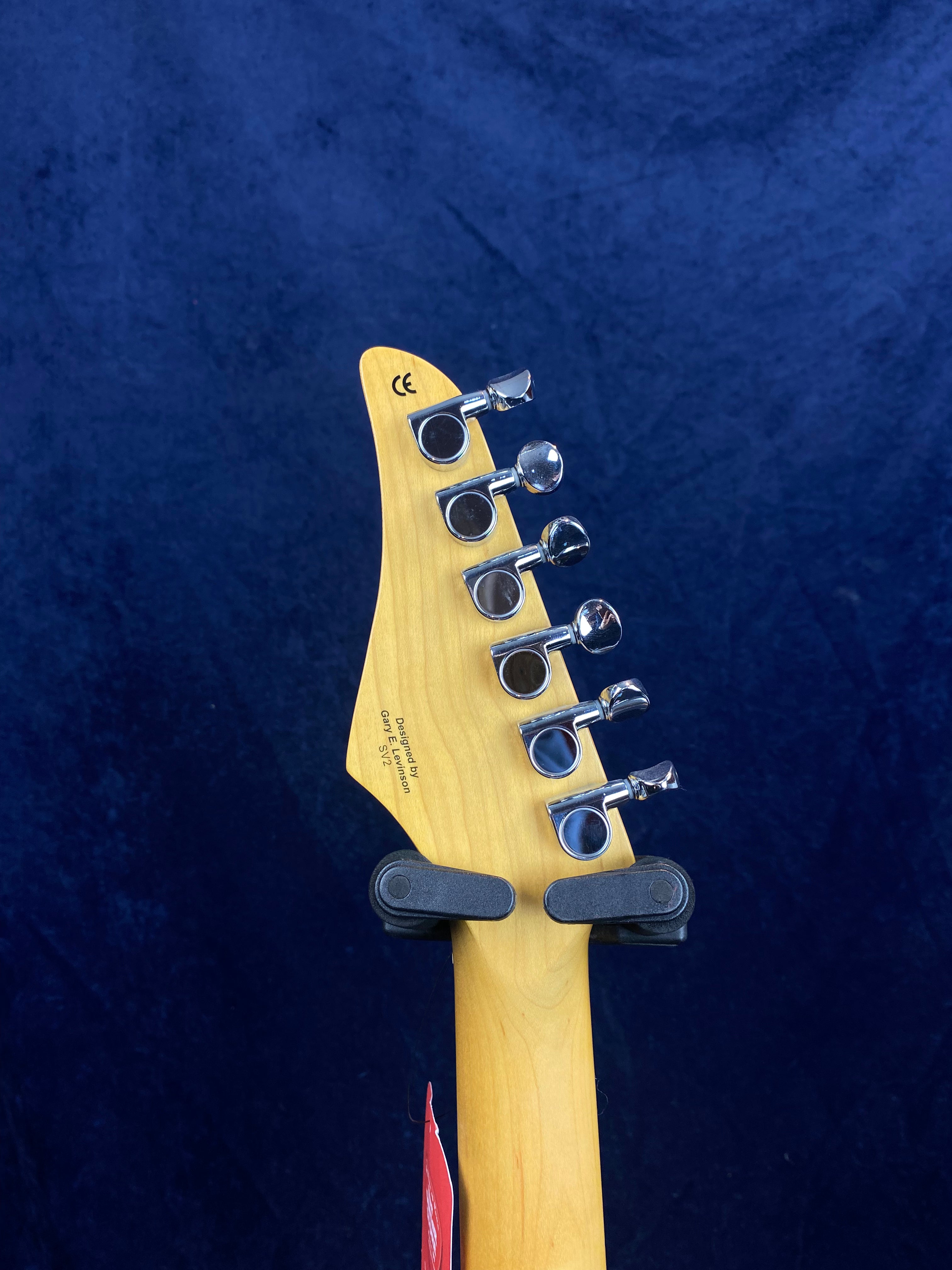 The back of an electric guitar headstock with six chrome tuning pegs, light wood finish, and small printed text near the top.