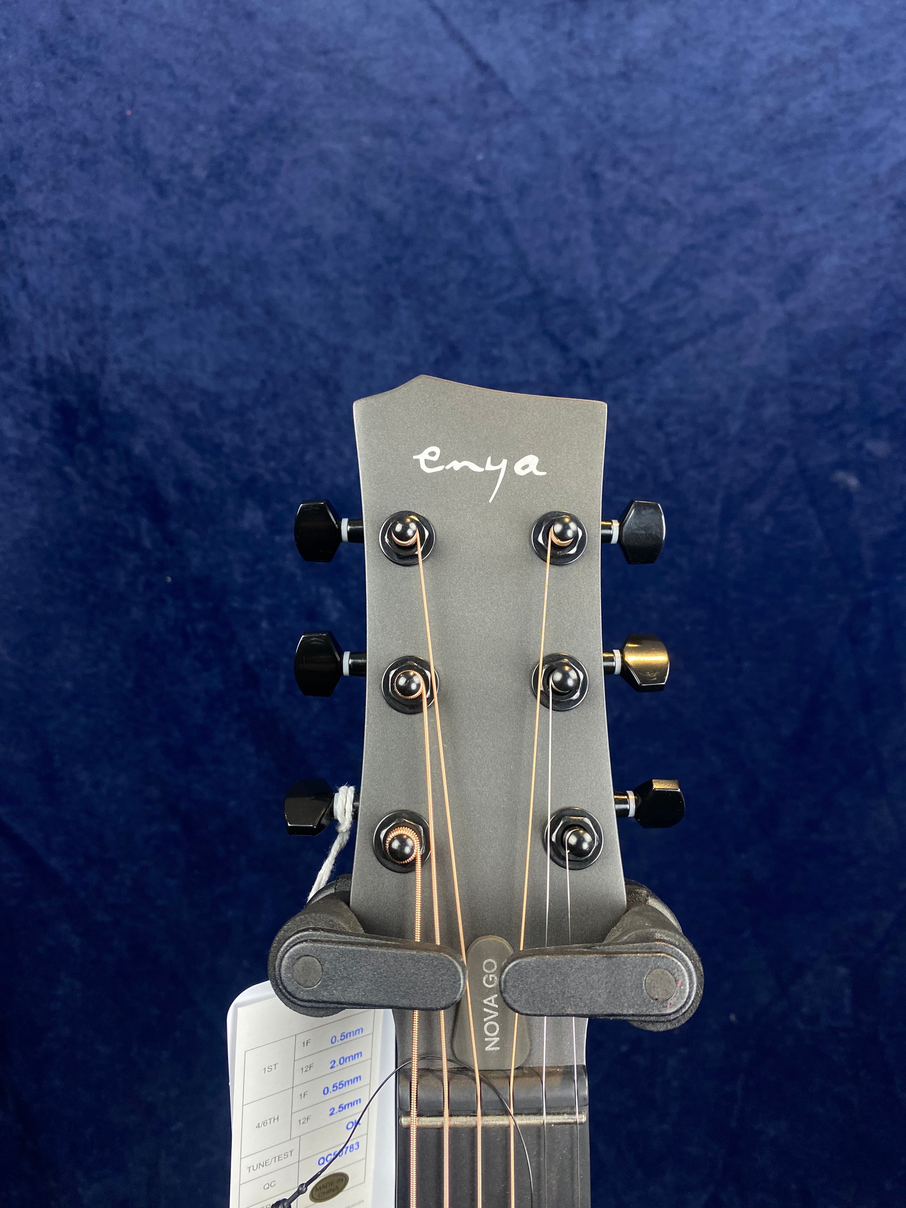 Gray Enya acoustic guitar headstock with six black tuning pegs, strings, and a tag attached, against a dark blue background.