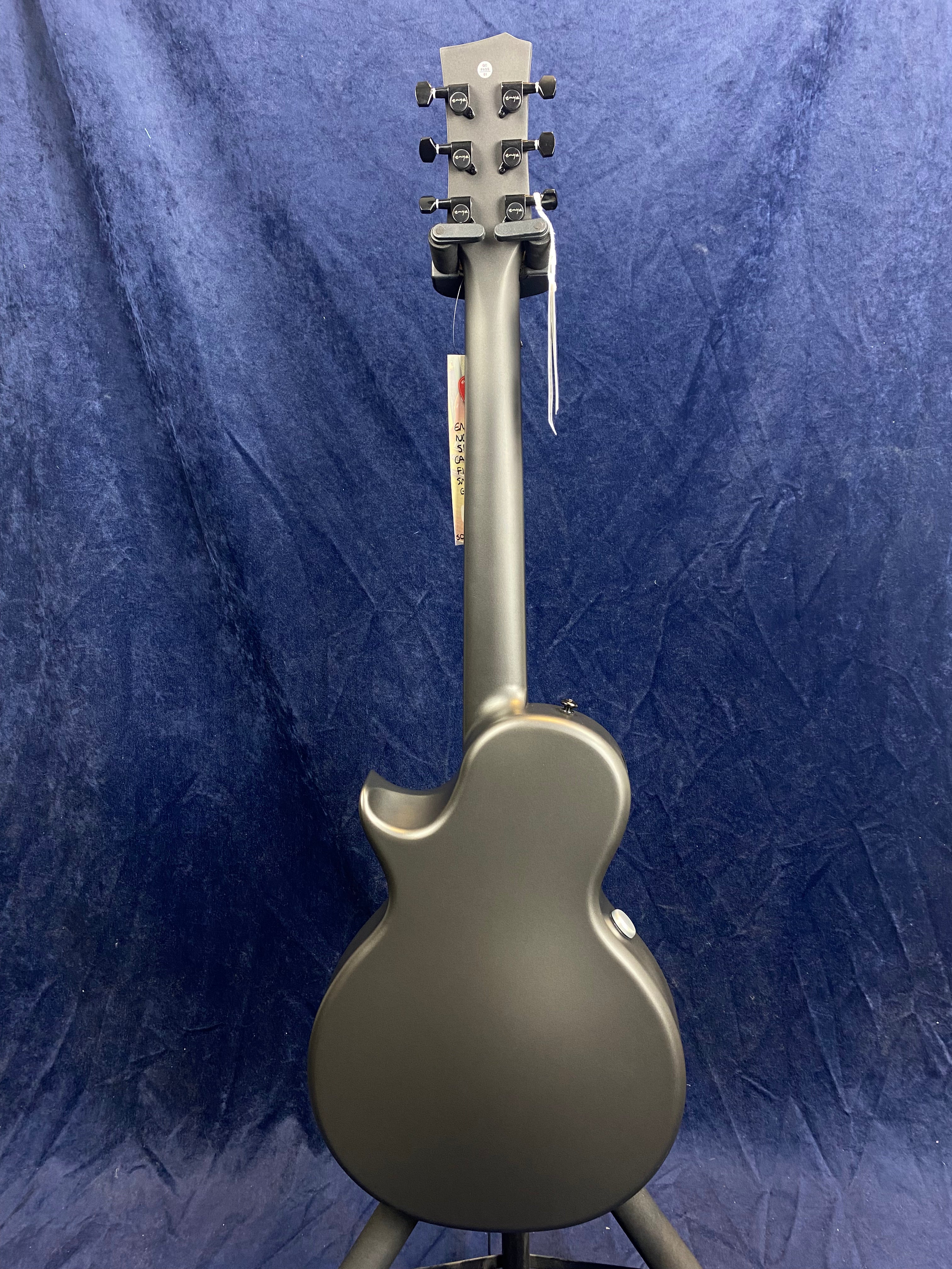 Matte black acoustic guitar shown from the back, single cutaway design, six tuning pegs, smooth finish, standing on a tripod against a dark blue background.