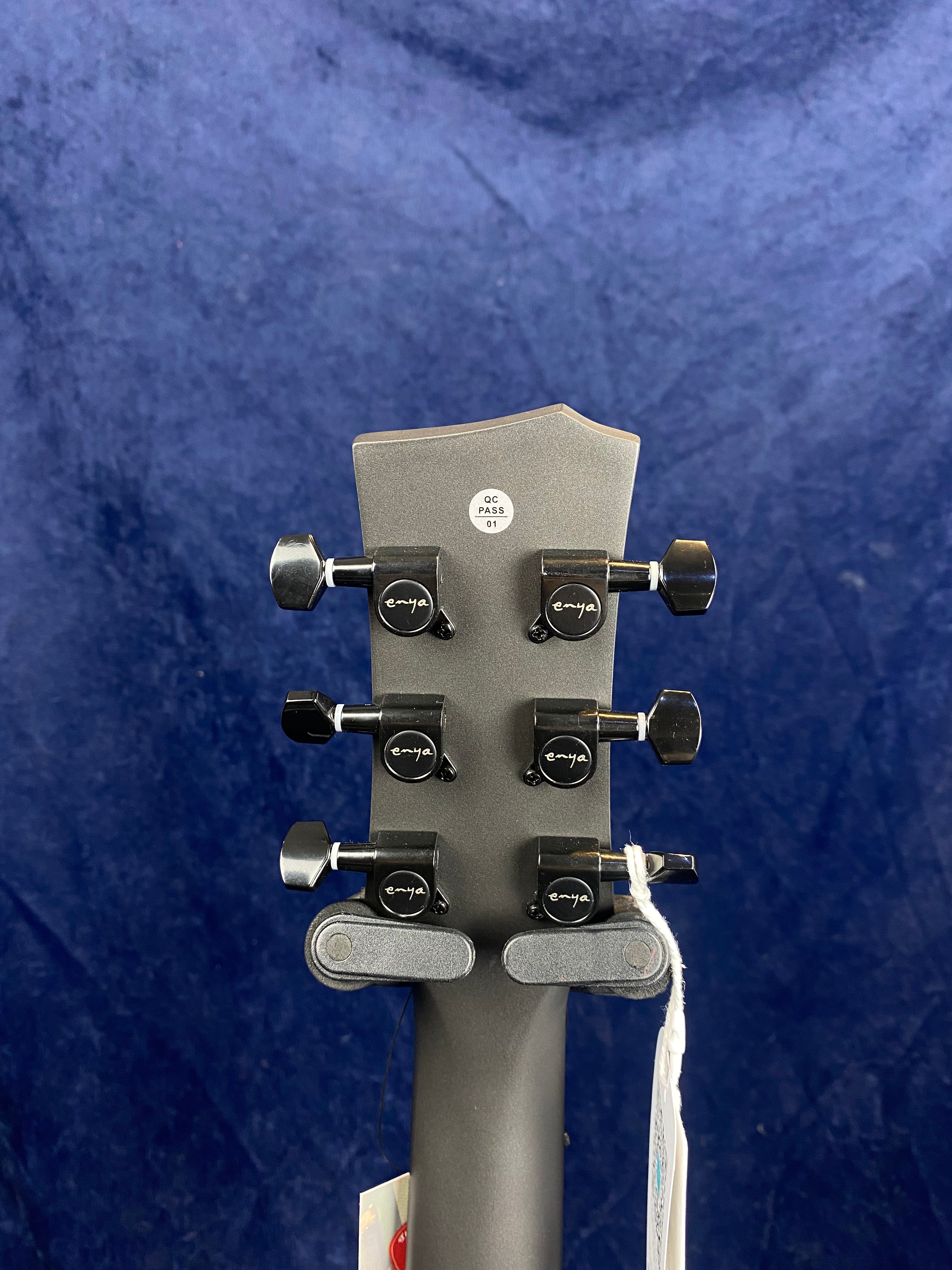 Six black tuning pegs on the back of a gray acoustic guitar headstock, with "enya" branding and a small QC PASS 01 sticker.