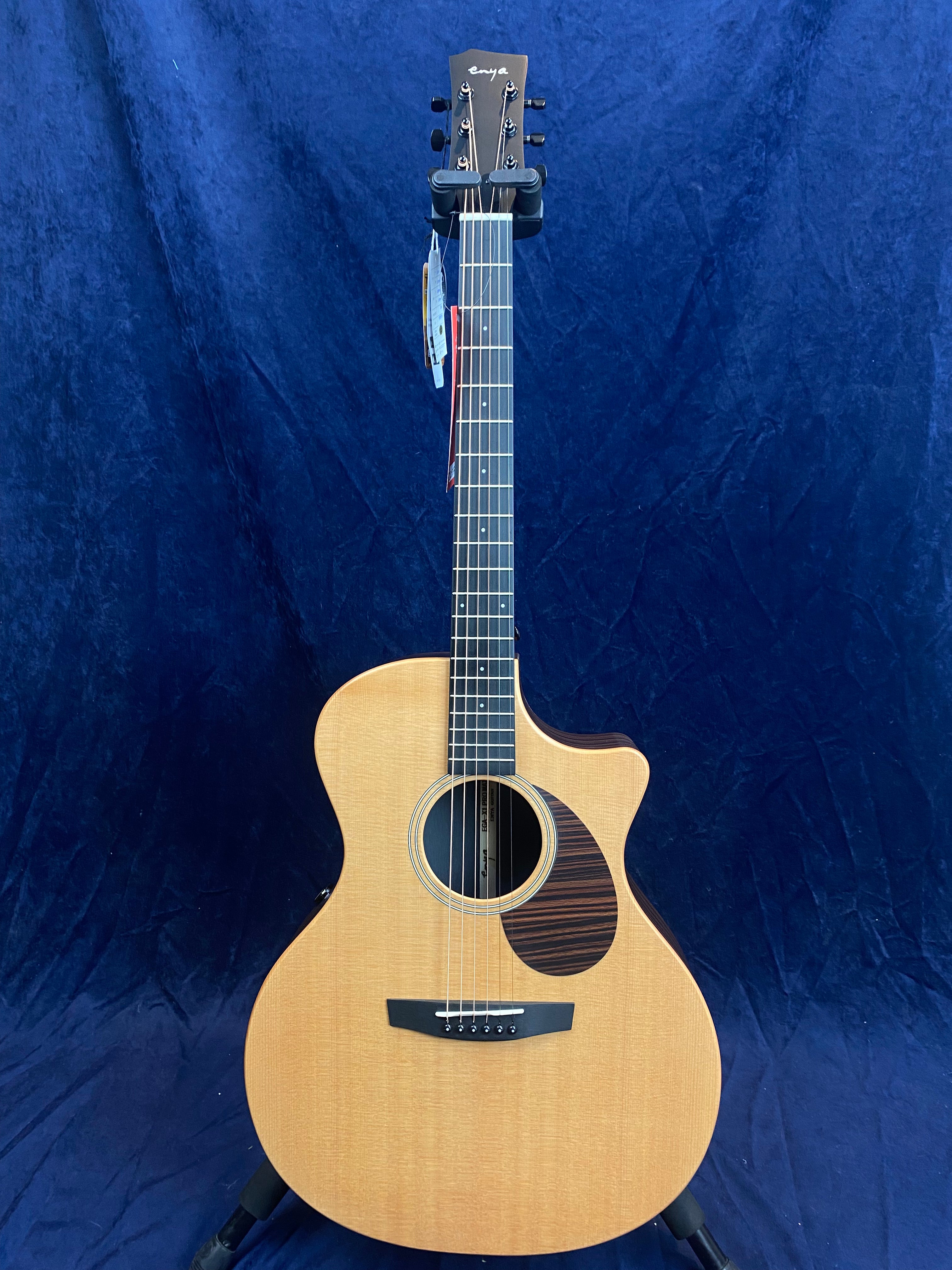 Light wood acoustic guitar with a single cutaway, dark wood pickguard, black bridge, and six tuning pegs, displayed upright against a blue backdrop.