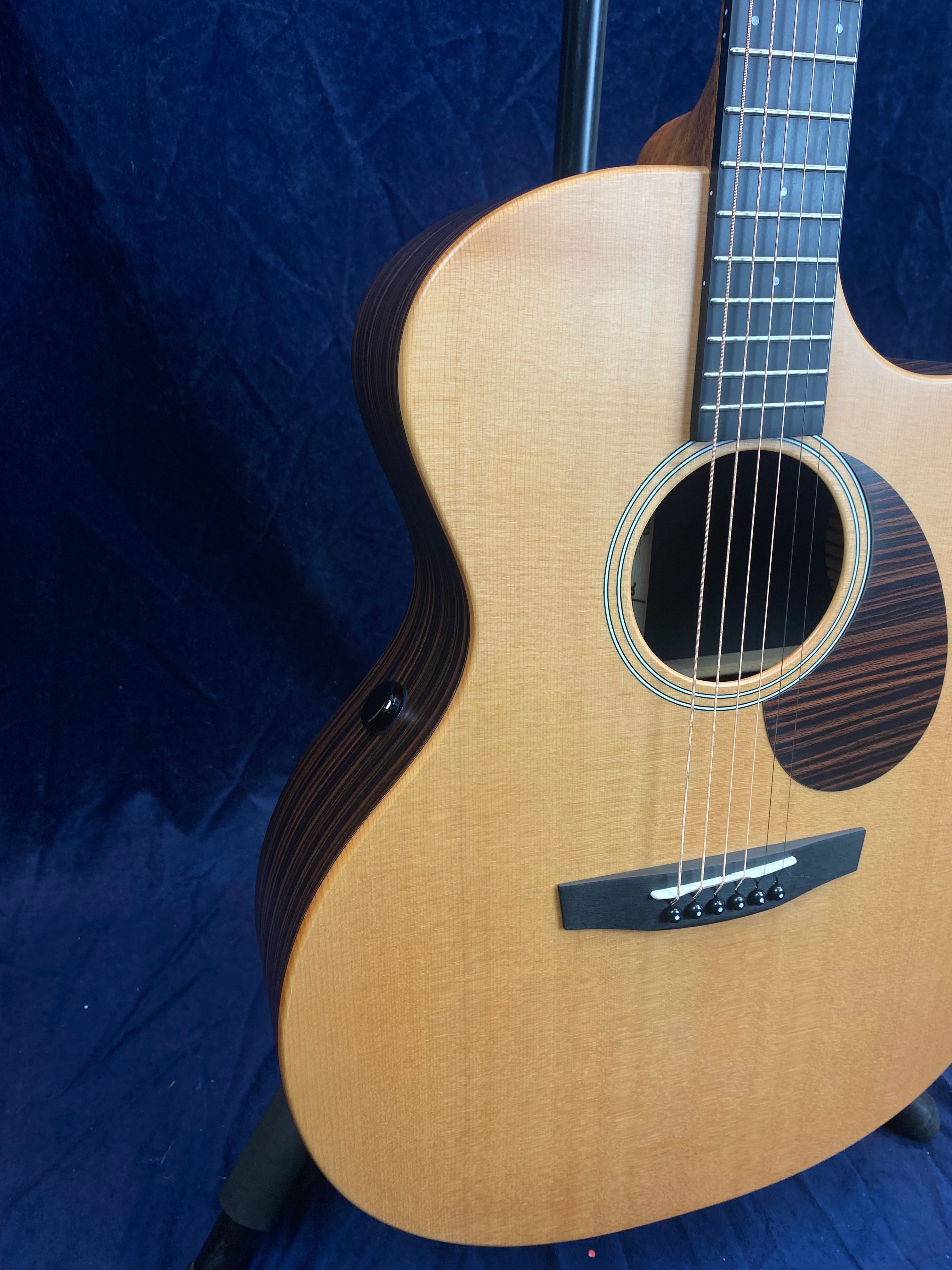 Light wood acoustic guitar with dark wood sides, circular sound hole, six strings, and dark bridge, shown on a stand against a dark blue background.
