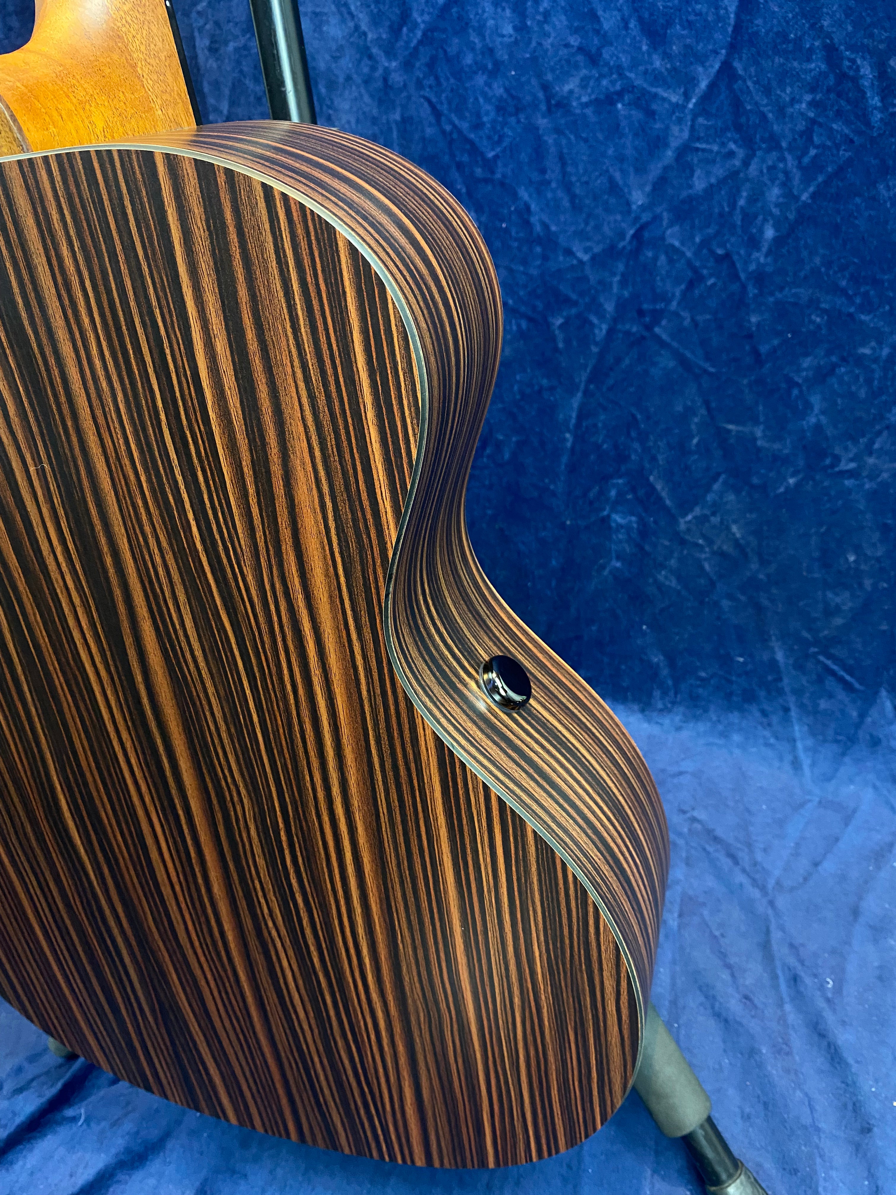 Acoustic guitar with dark brown and black striped wood body, glossy finish, and a black strap button, shown against a deep blue textured background.