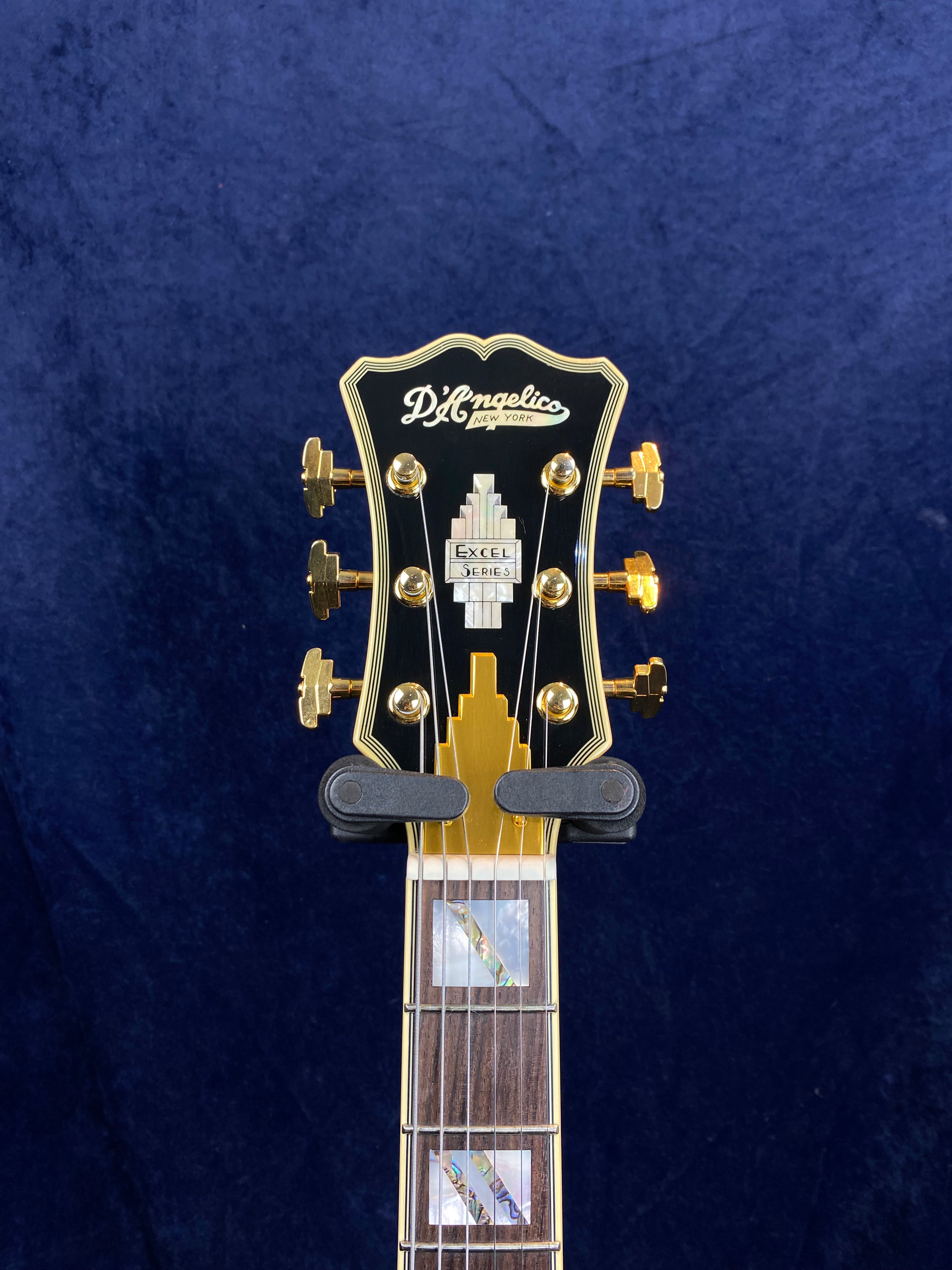 D’Angelico Excel Series electric guitar headstock with gold tuners, black faceplate, art deco inlay, and part of the fretboard visible.