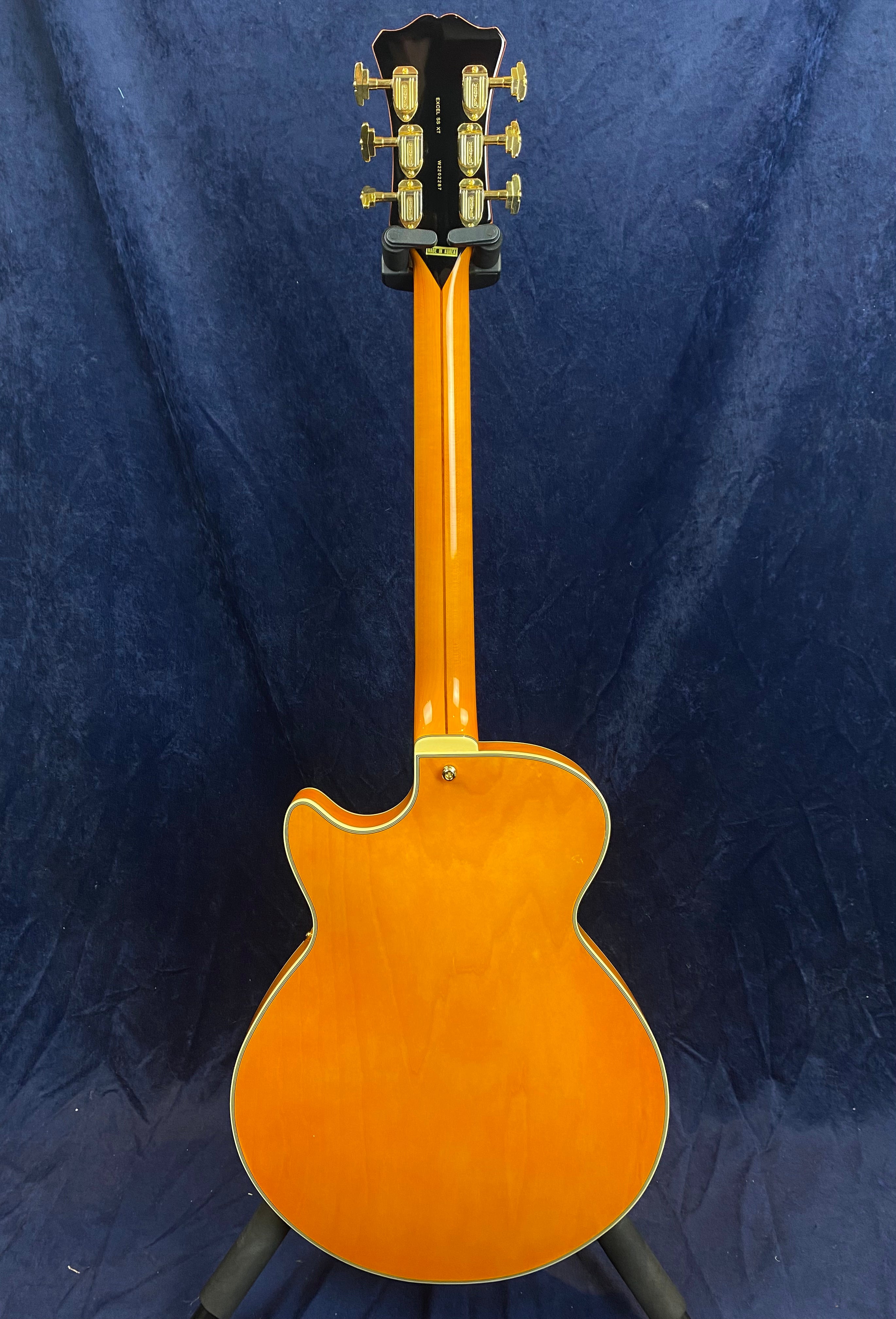 Sunburst orange electric guitar shown from the back, single cutaway body, gold tuning pegs, cream binding, displayed on a stand against a dark background.