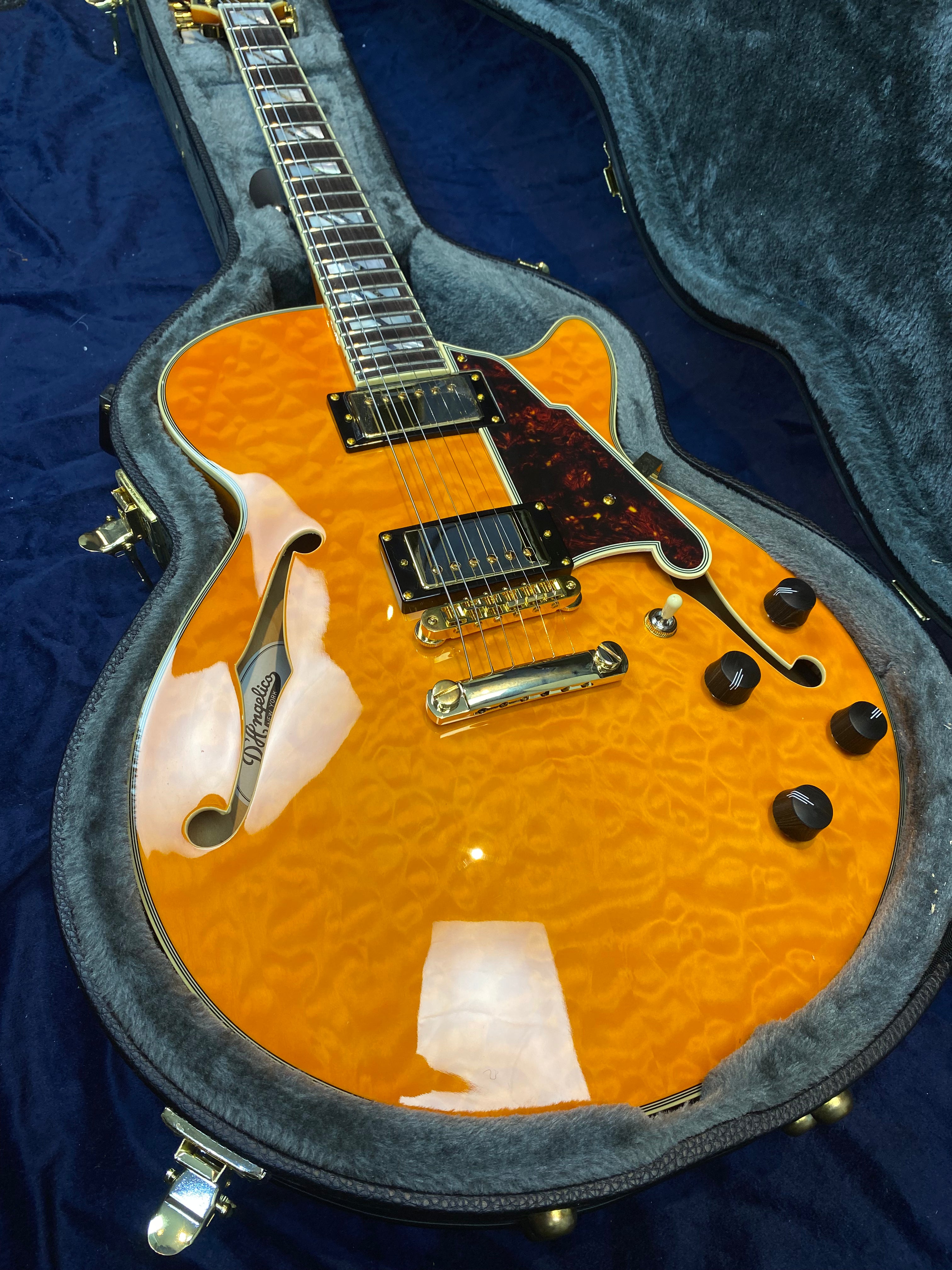Electric guitar with a bright amber quilted maple top, gold hardware, black knobs, and a tortoiseshell pickguard, resting in a gray plush case.