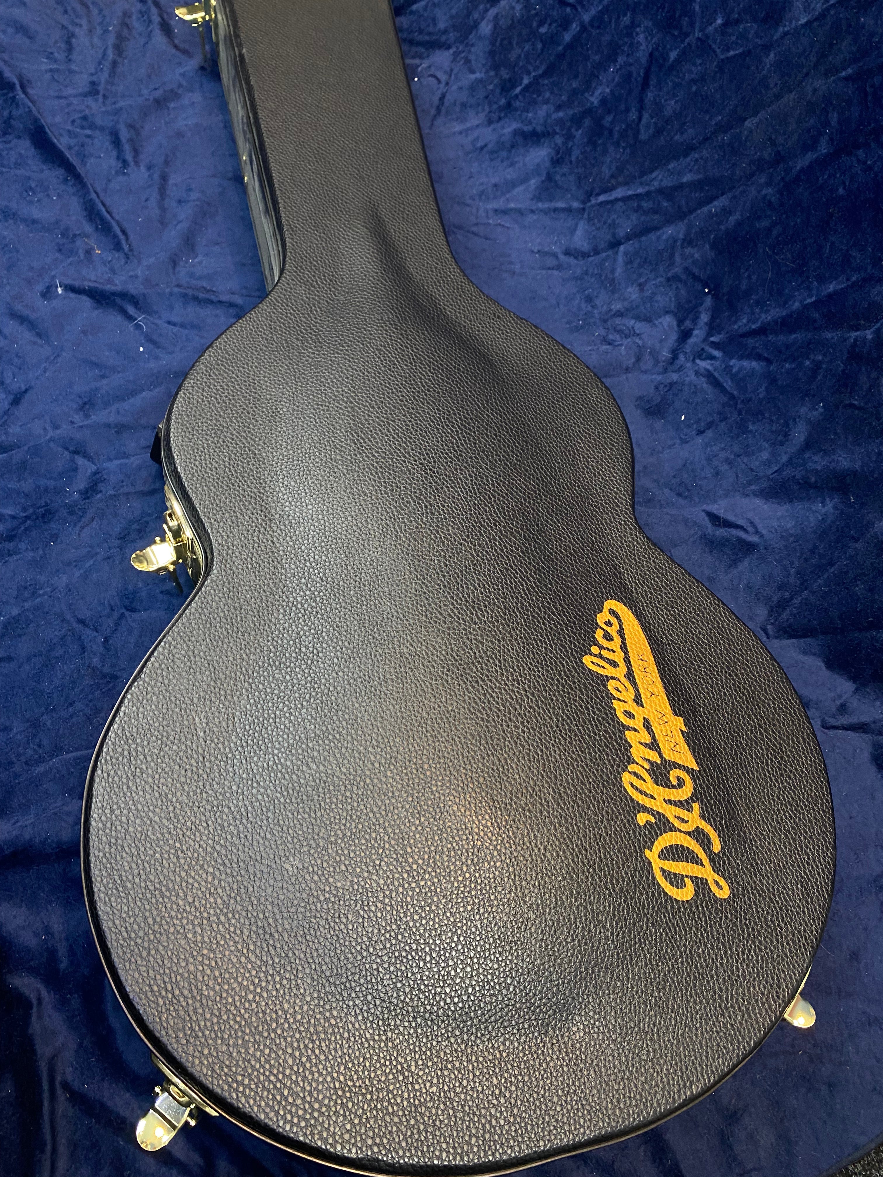 Black textured hardshell electric guitar case with gold D’Angelico script logo, resting on dark blue fabric, gold latches visible on the sides.