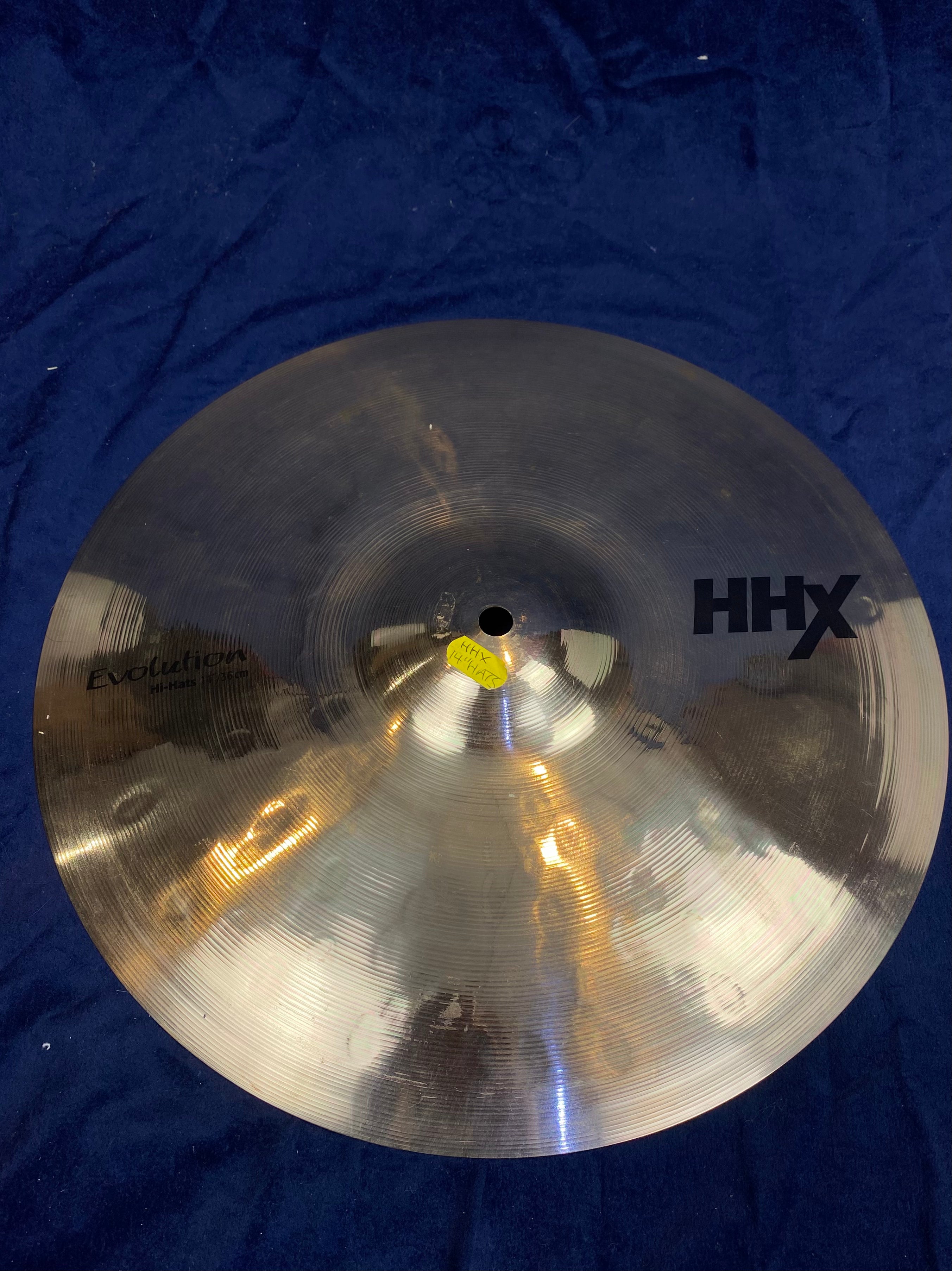 Shiny metal hi-hat cymbal labeled HHX Evolution, 14-inch size, resting on a dark blue cloth background.