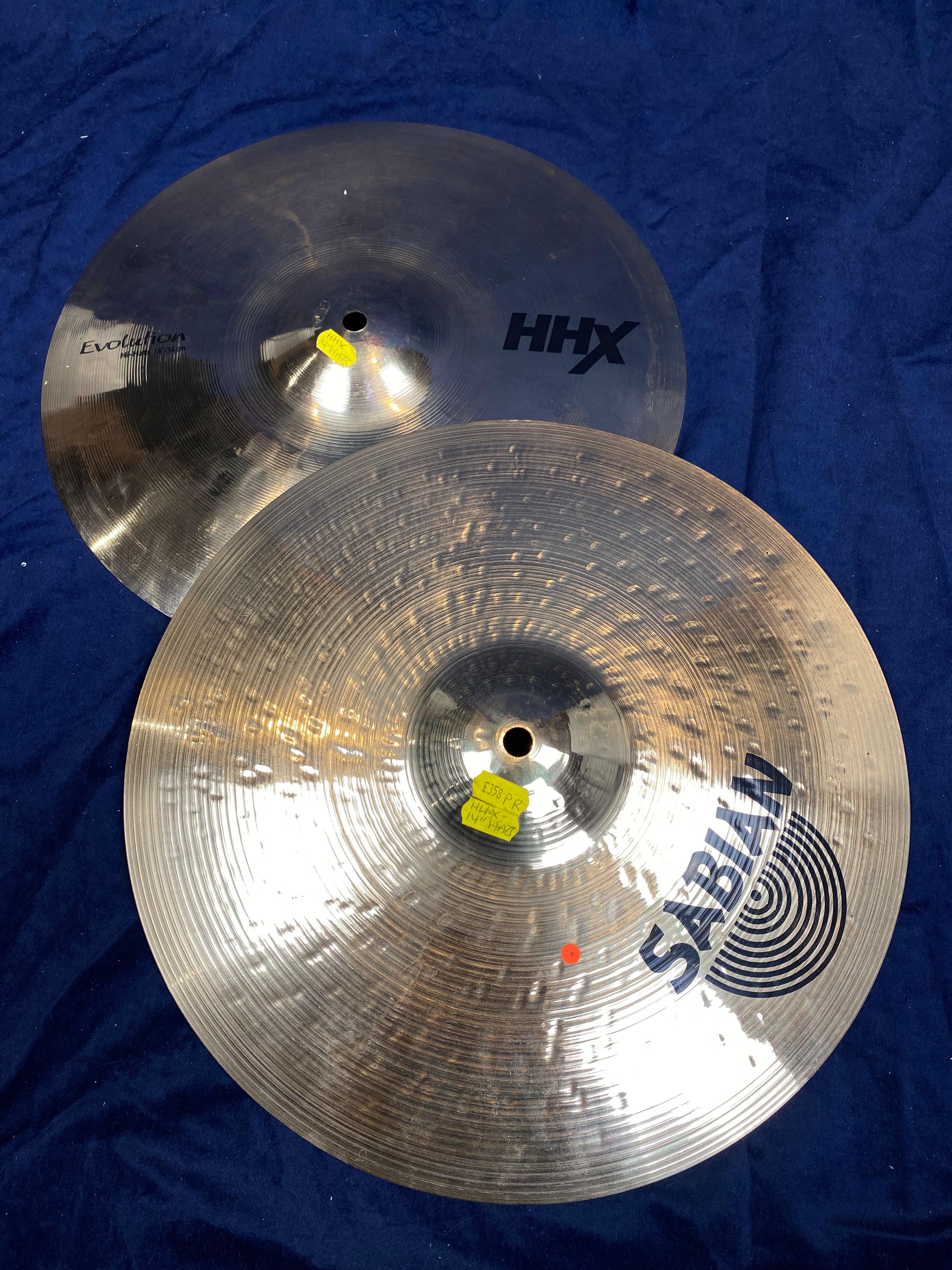 Two shiny metal cymbals with "SABIAN" and "HHX" logos rest on a dark blue cloth, each with a small yellow sticker near the center hole.