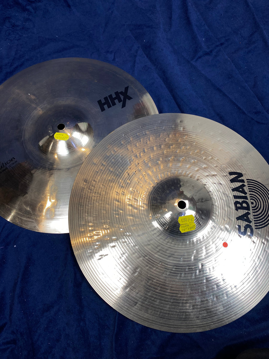 Two shiny metal cymbals labeled HHX and Sabian rest on a dark blue fabric, used for drums and percussion setups.
