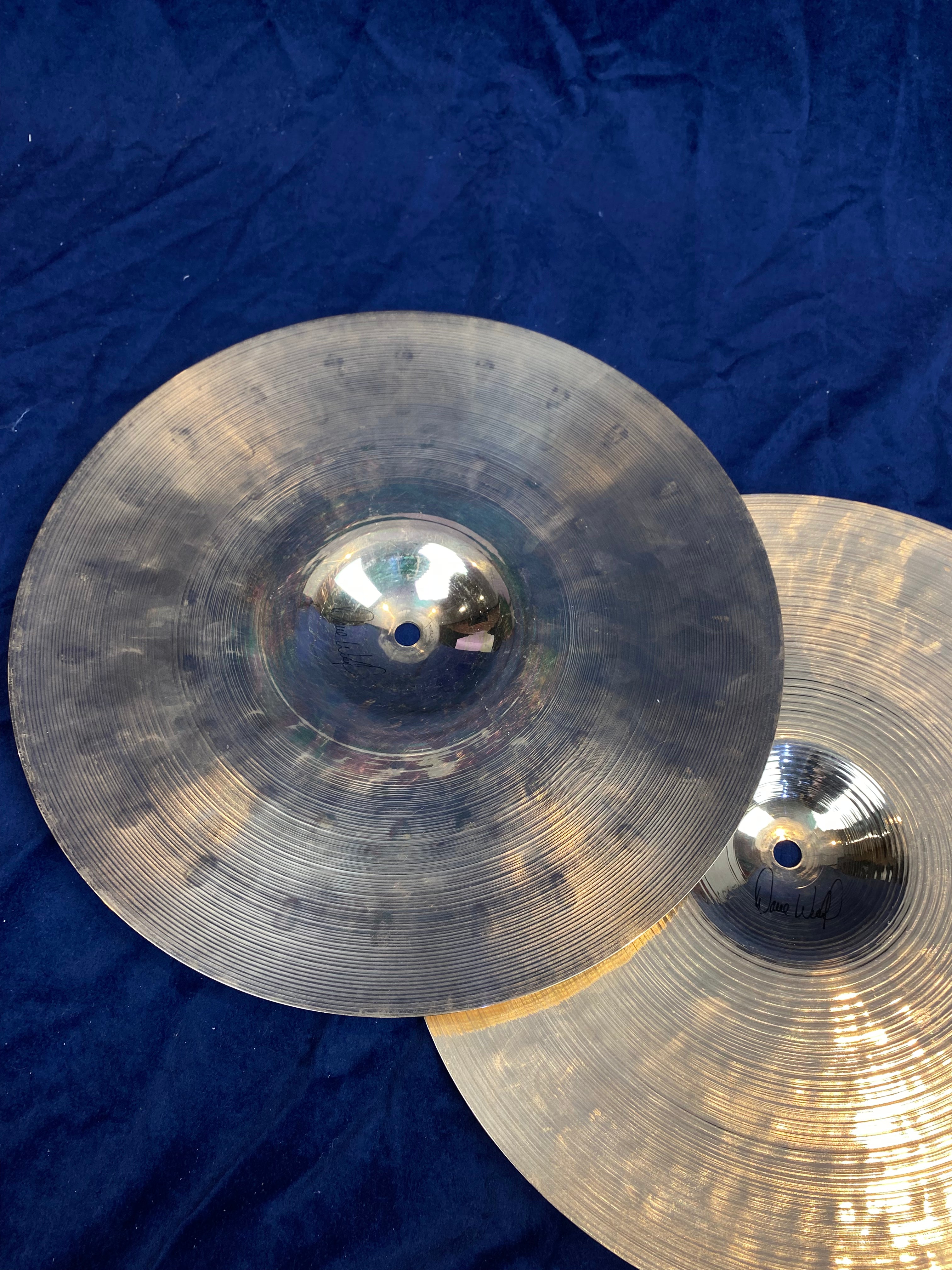 Two metallic cymbals with concentric grooves, one tarnished and darkened, the other shiny with a visible signature, lying on a dark blue fabric.