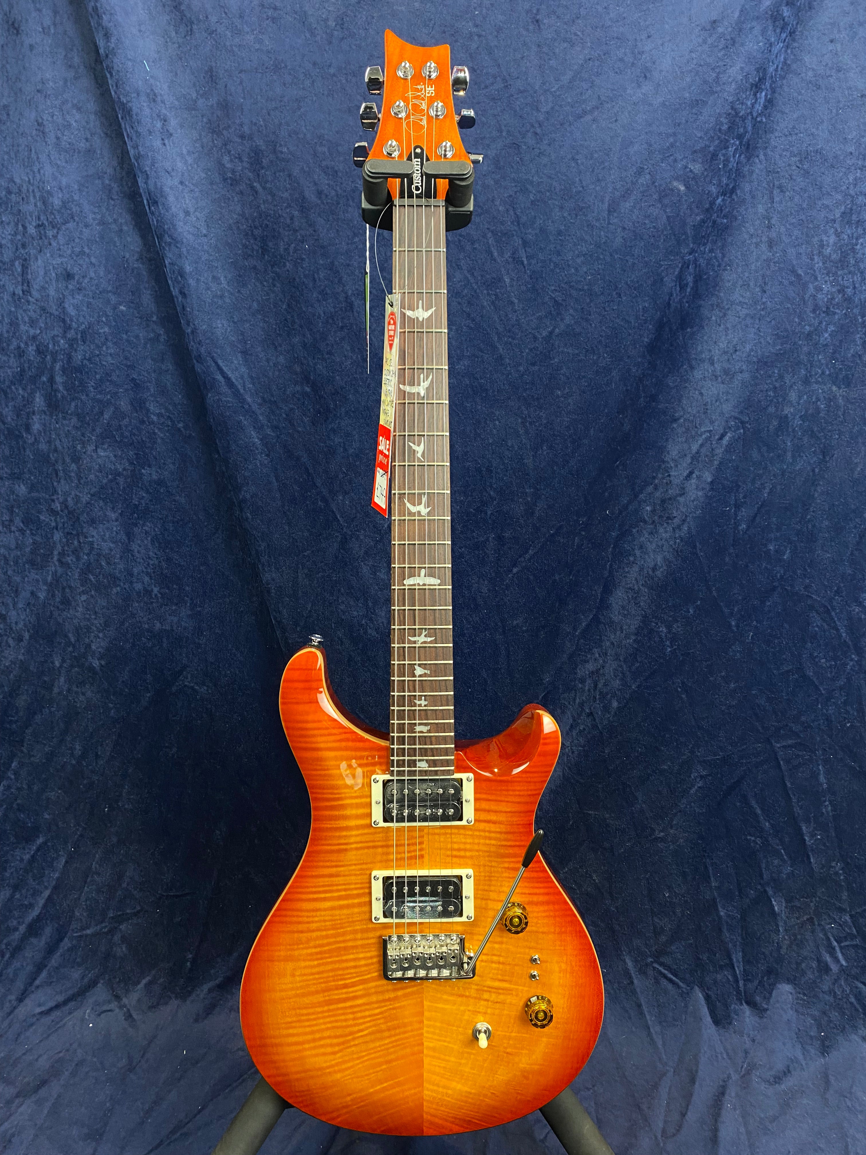 Electric guitar with a glossy orange sunburst finish, bird-shaped inlays on the fretboard, two pickups, and three control knobs, standing against a dark background.