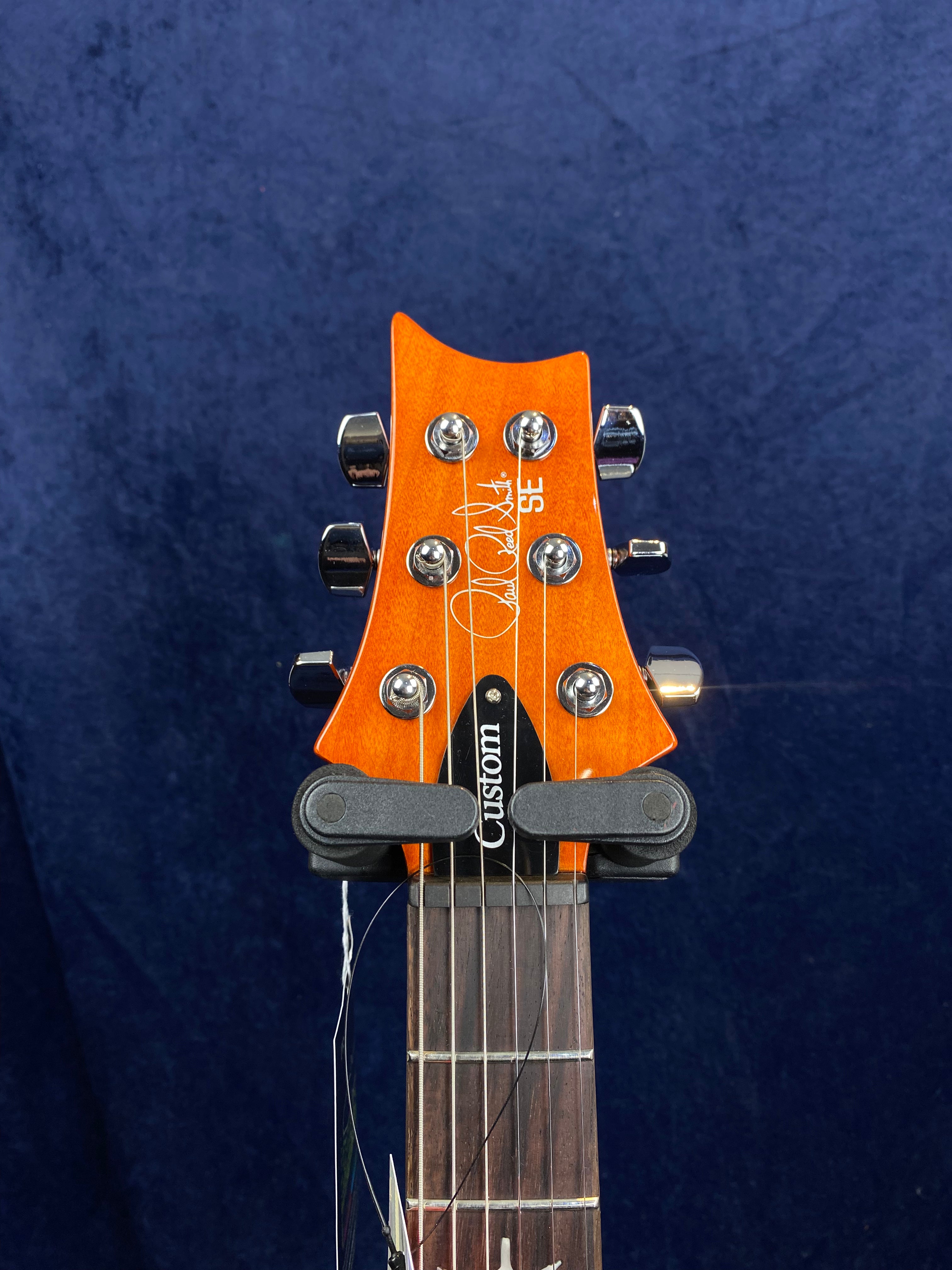 Orange electric guitar headstock with six chrome tuning pegs, Custom truss rod cover, and white Paul Reed Smith SE signature.