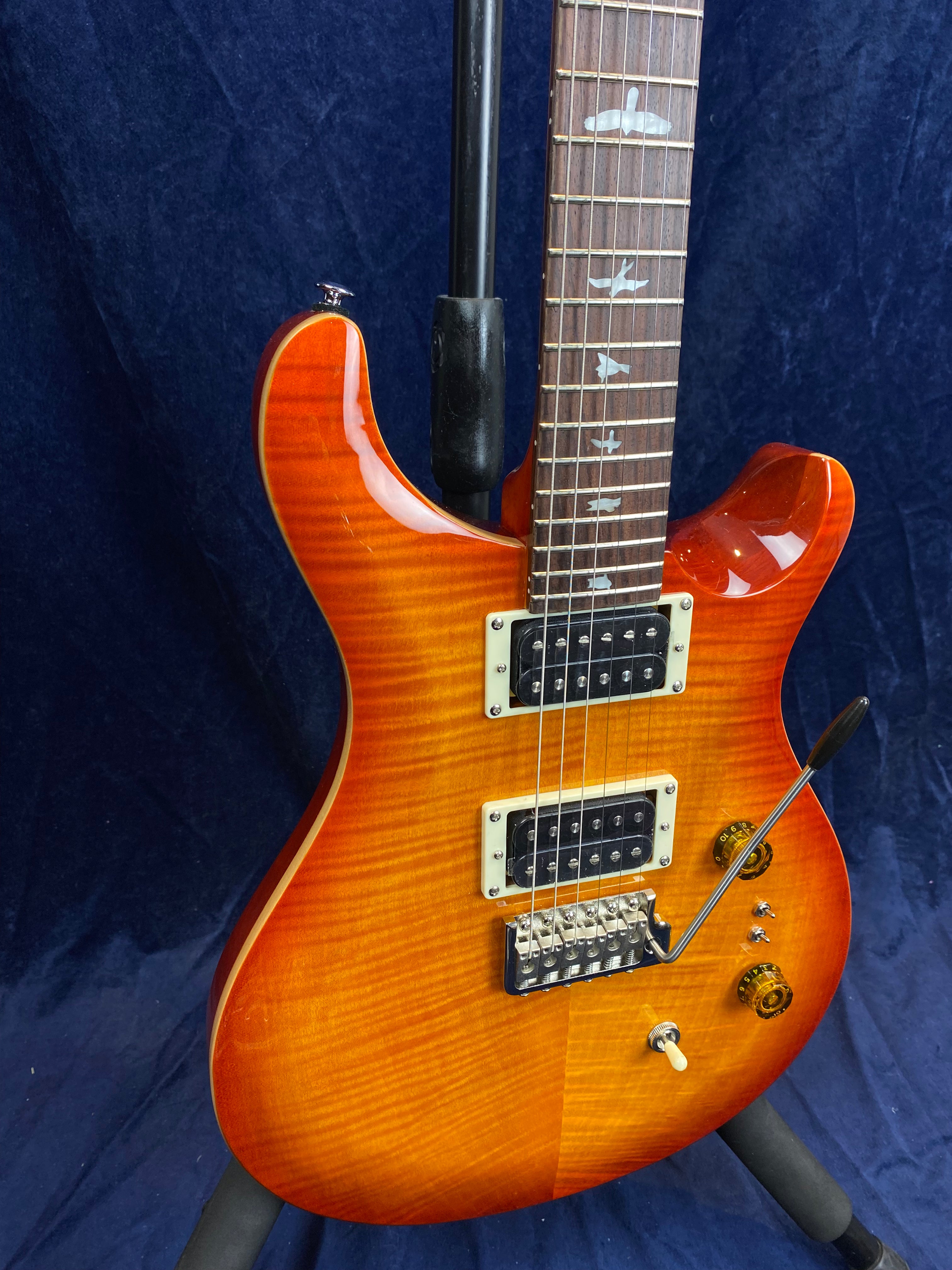 Electric guitar with a glossy orange sunburst finish, two humbucker pickups, bird inlays on the fretboard, and a whammy bar near the bridge.