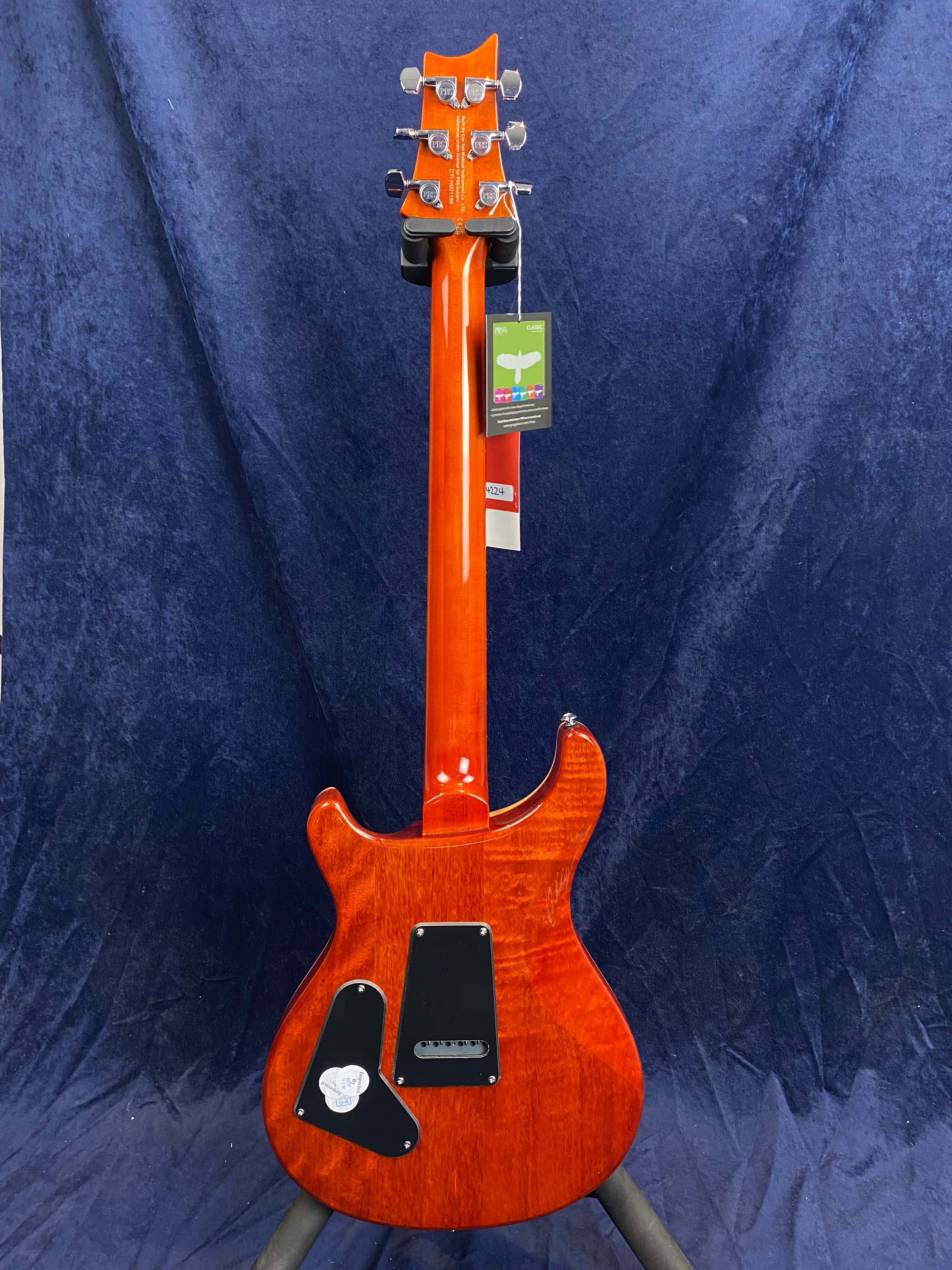 Electric guitar with a glossy red-orange finish, six tuning pegs, and two black plastic covers on the back; tags hang from the headstock.