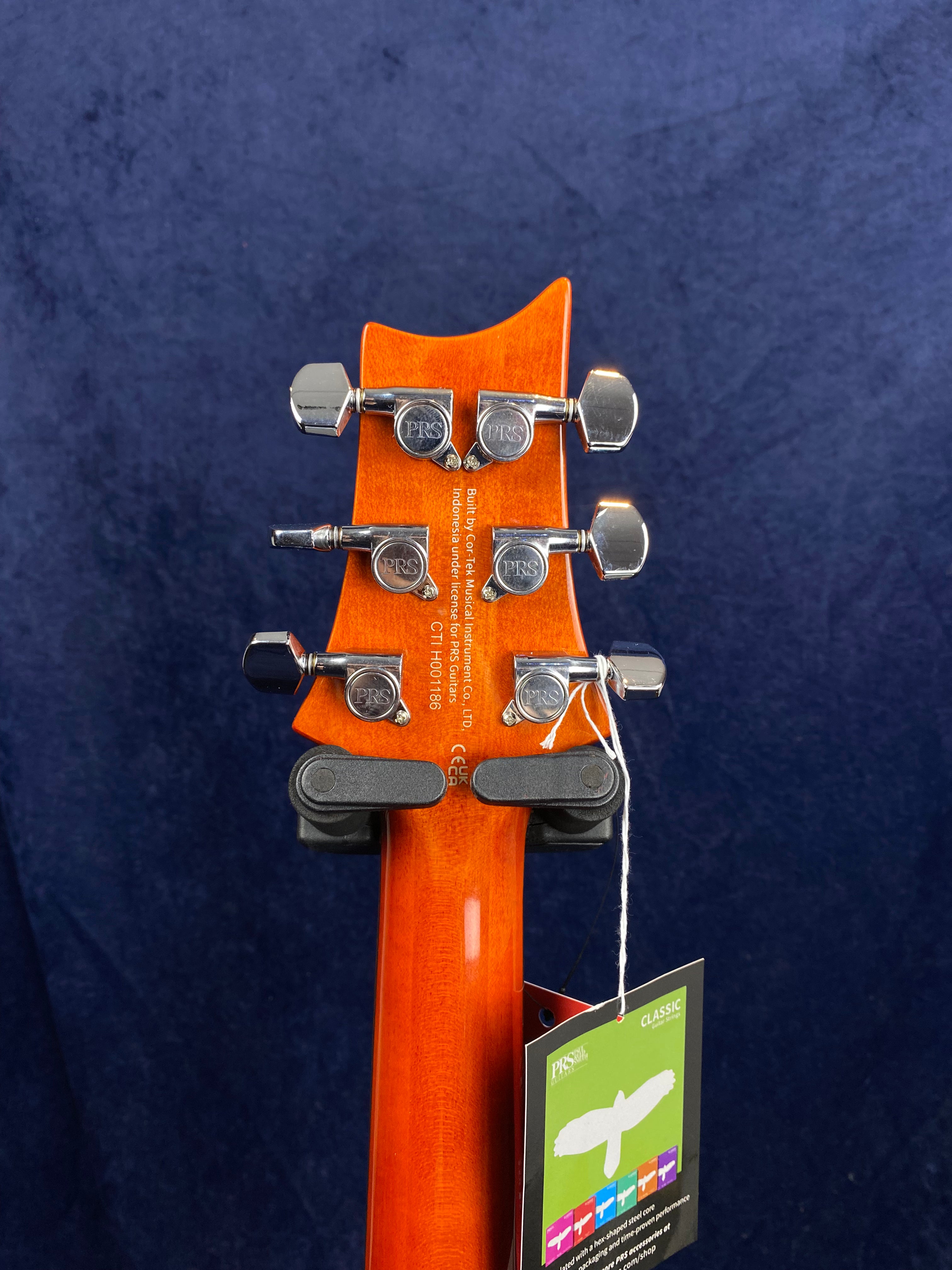Six chrome PRS tuning pegs on a glossy orange electric guitar headstock, with a product tag hanging by a string.