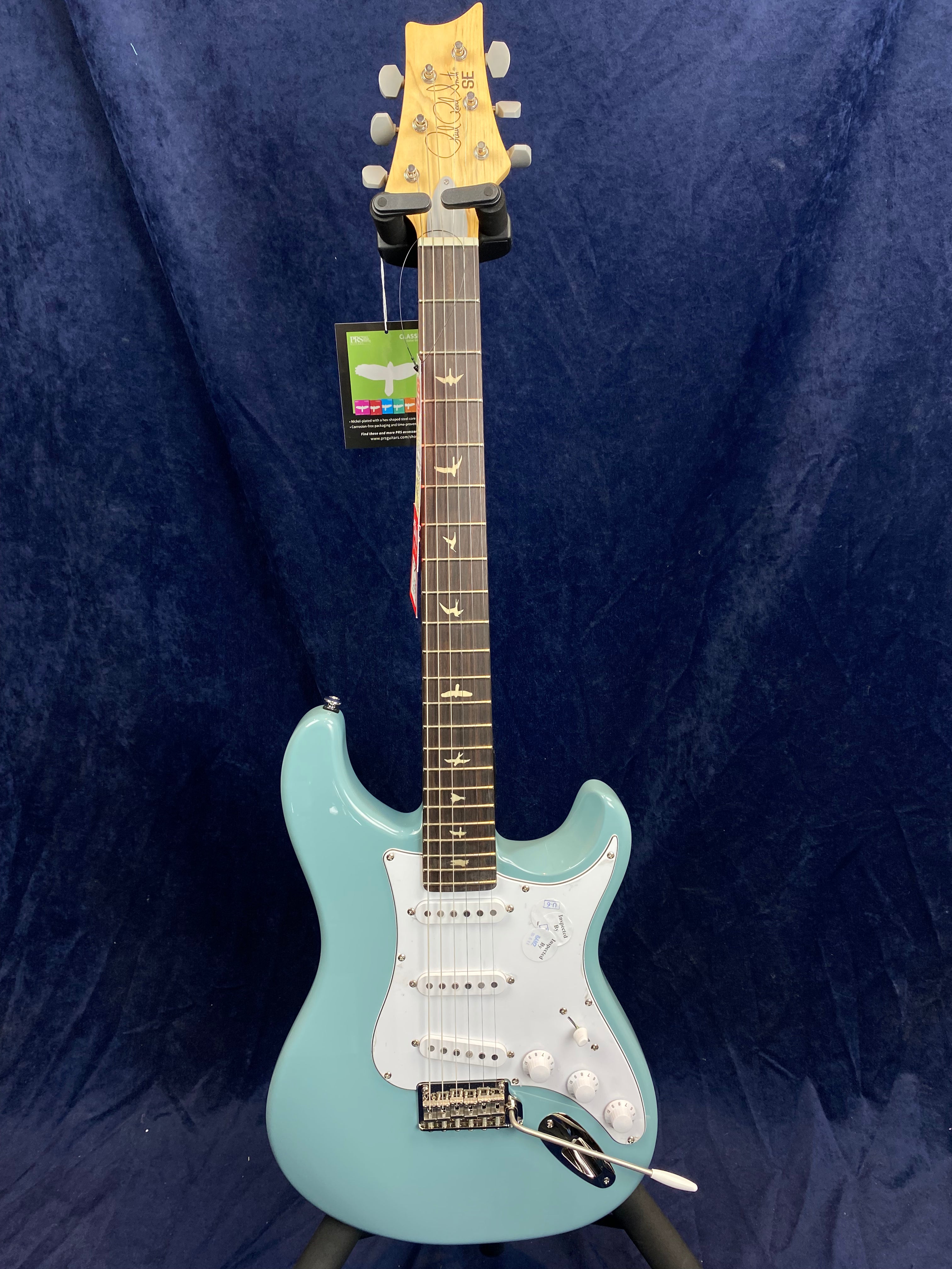 Light blue electric guitar with a white pickguard, three single-coil pickups, bird inlays on the fretboard, and a maple headstock marked "PRS SE."