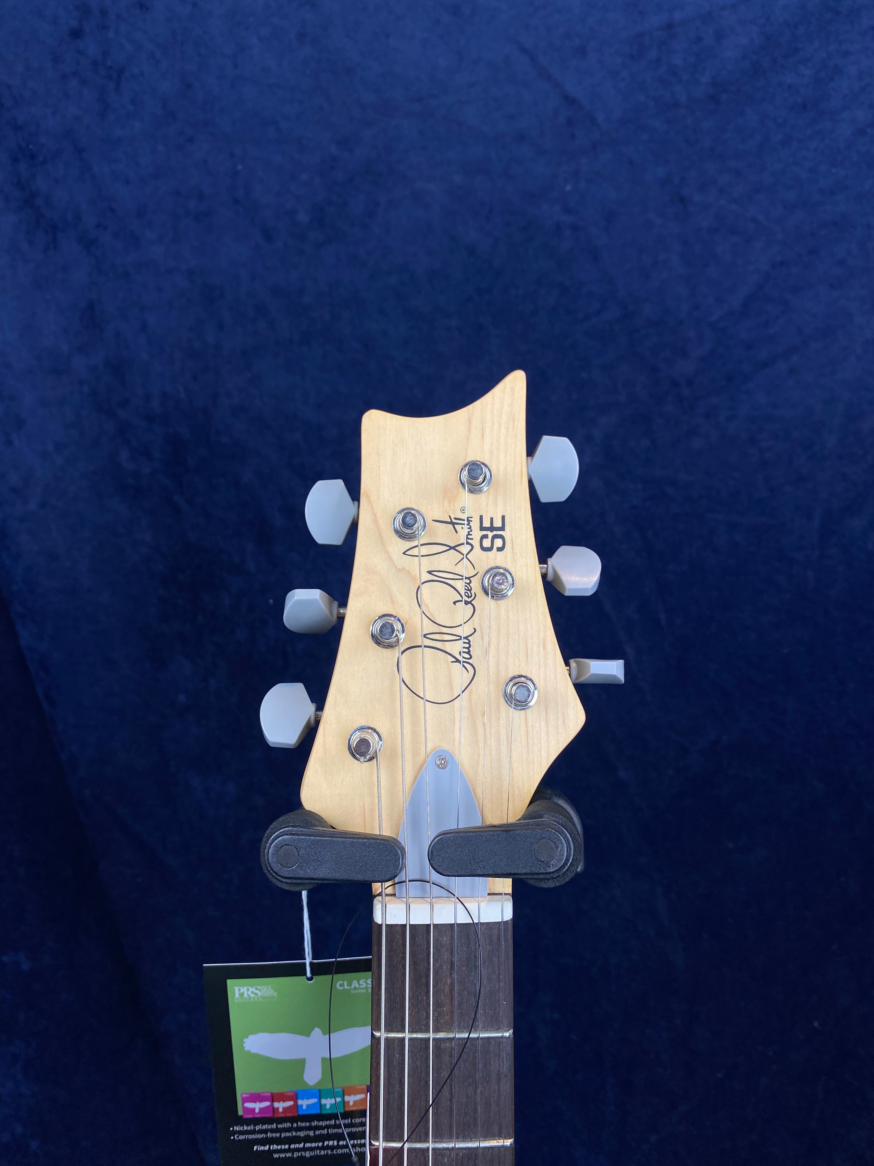 Electric guitar headstock with six tuning pegs, "SE" and signature printed on light wood, and a tag hanging from the neck.