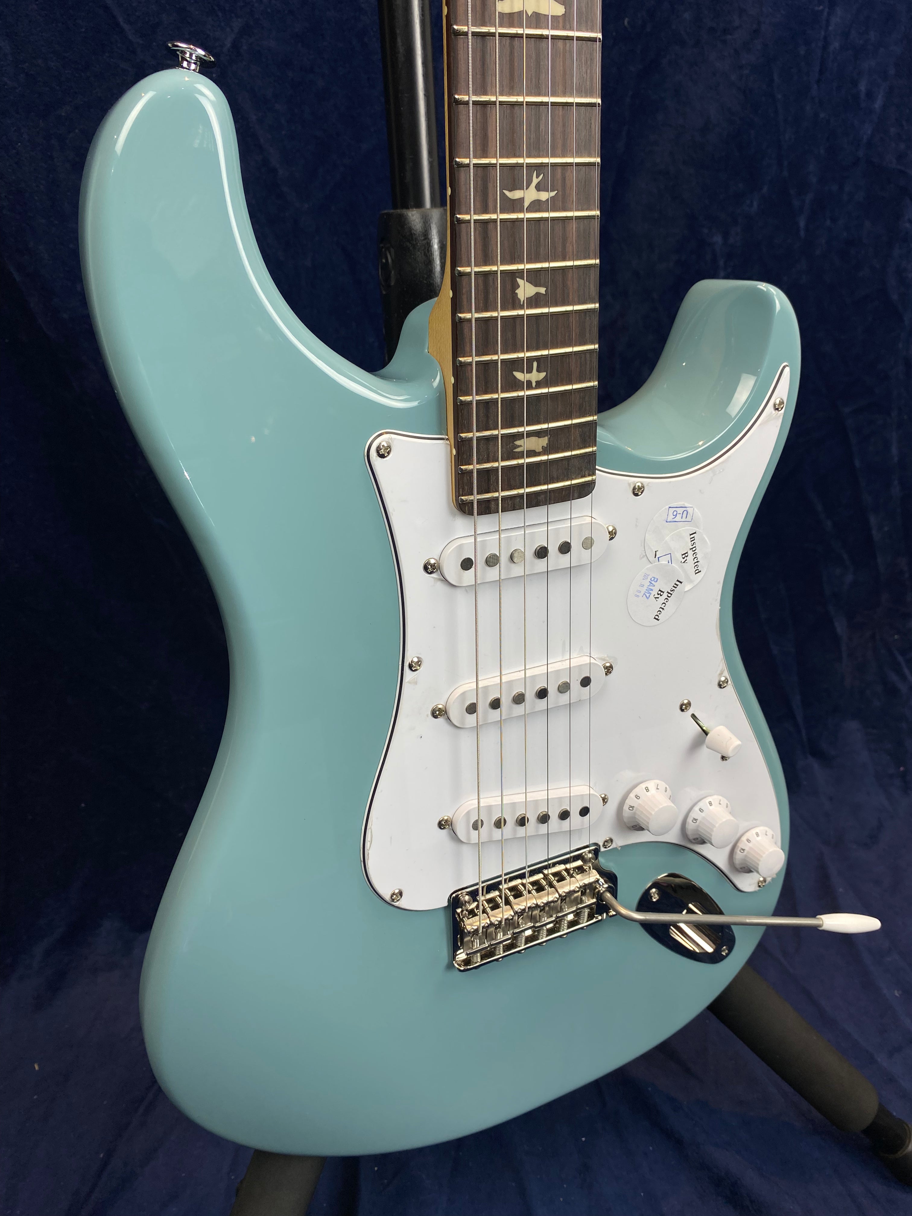 Light blue electric guitar with a white pickguard, three single-coil pickups, tremolo bar, and bird inlays on the dark fretboard.