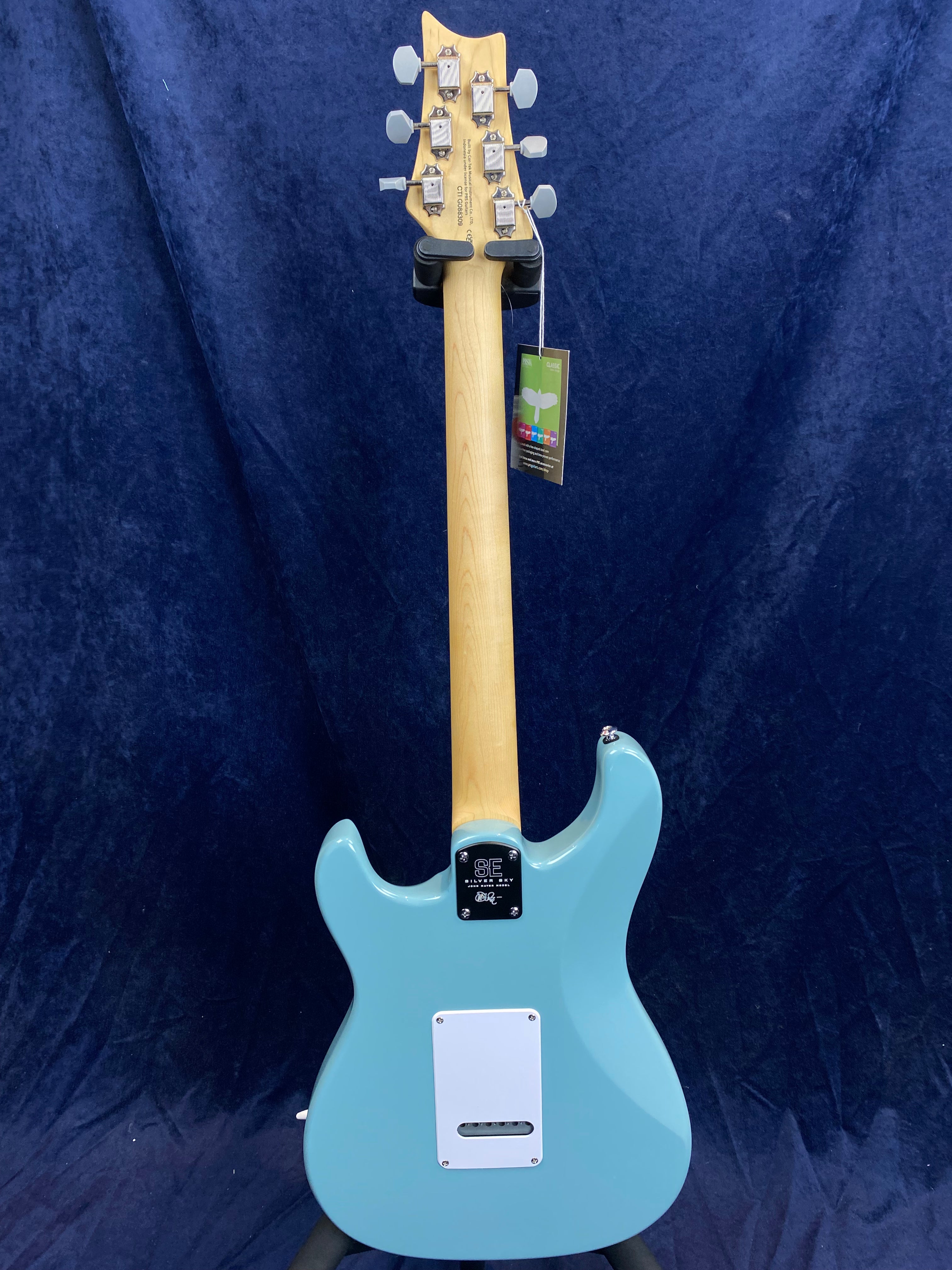 Light blue electric guitar shown from the back, with six tuning pegs, a light wood neck, white electronics cover, and a tag hanging from a tuner.