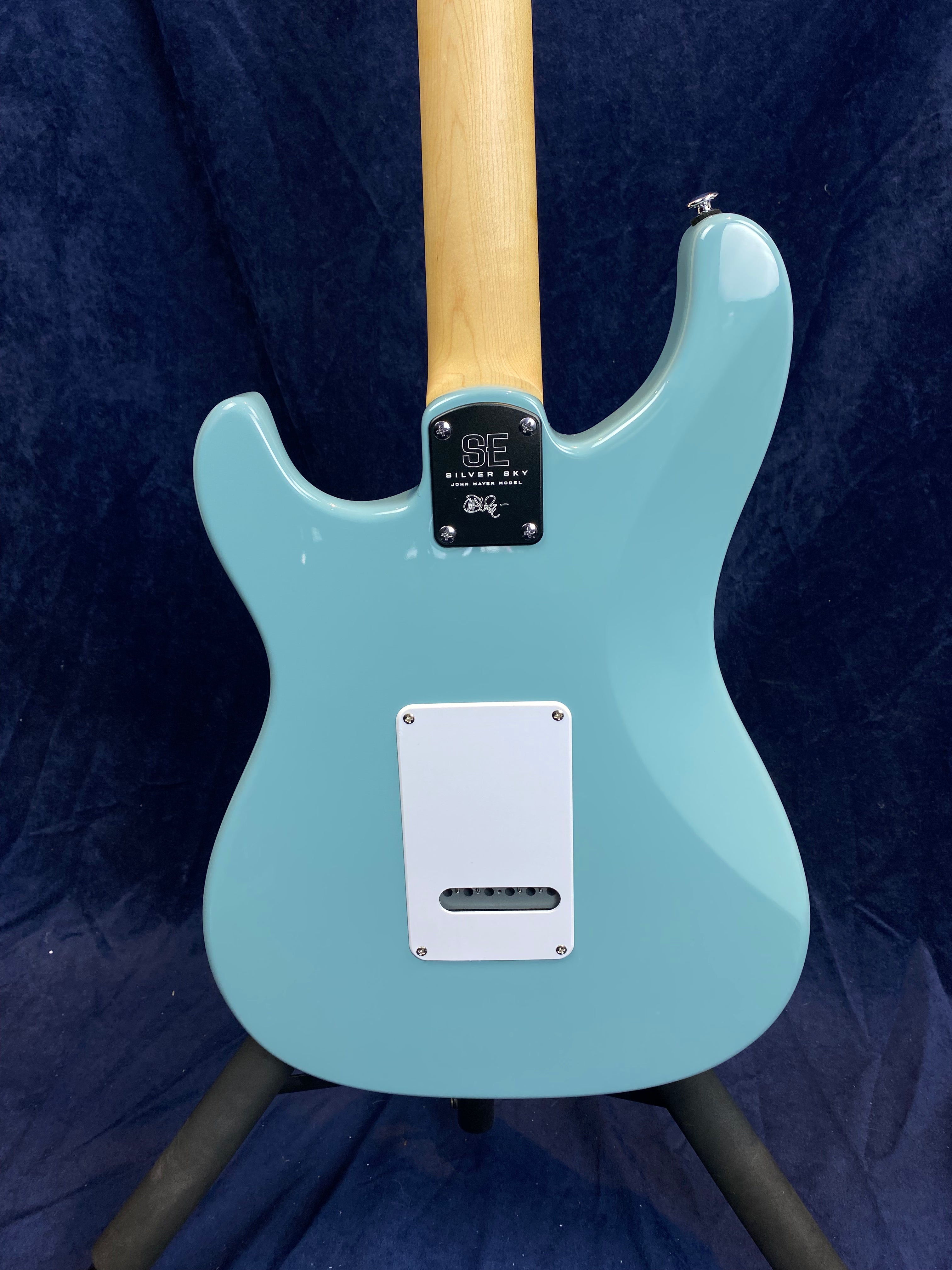 Light blue electric guitar seen from the back, natural wood neck, white tremolo cover, "SE Silver Sky" branding on neck plate, dark blue background.