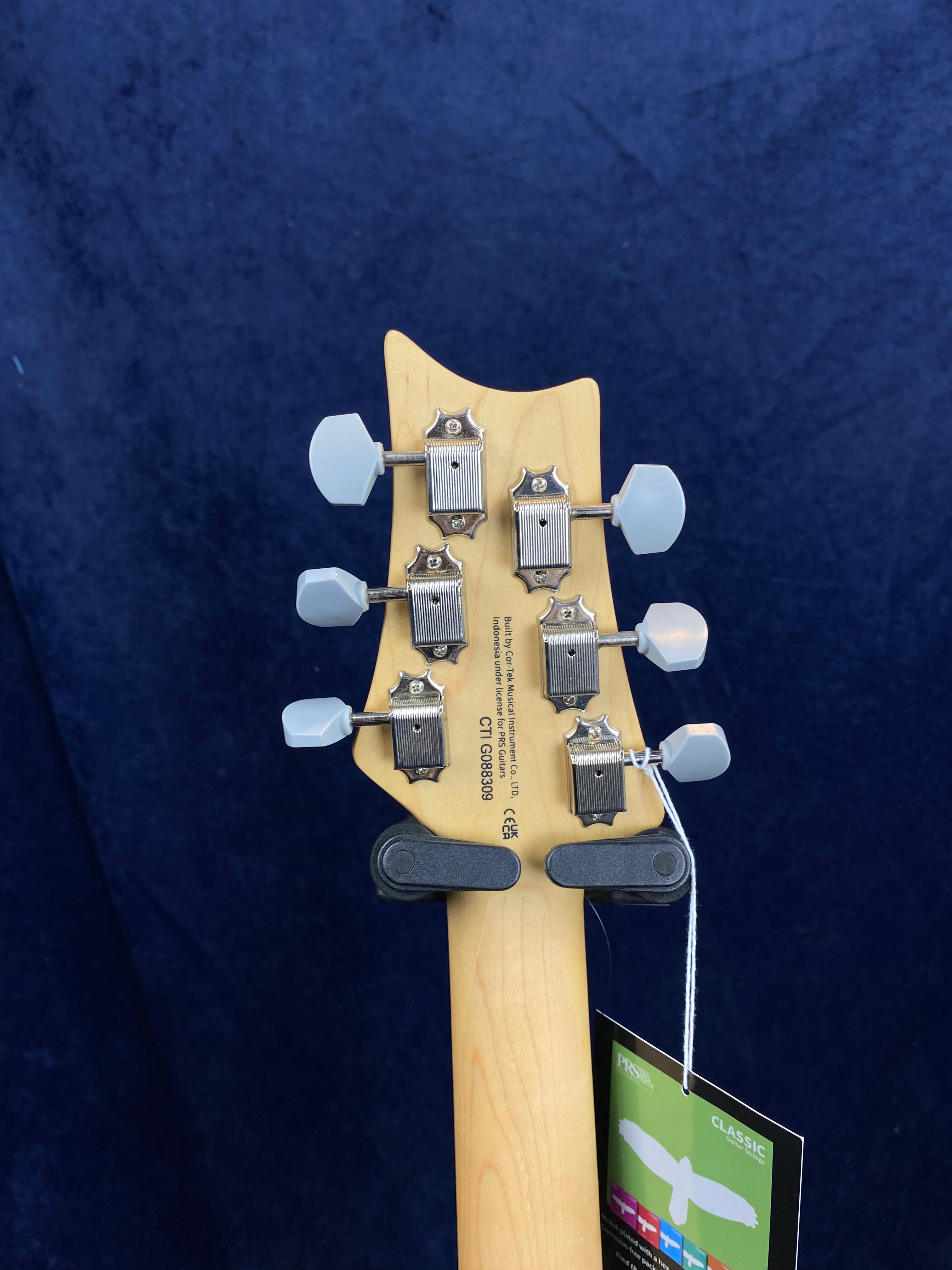 Six tuning pegs with white knobs on a light wood electric guitar headstock, seen from the back, with a tag hanging from one tuner.