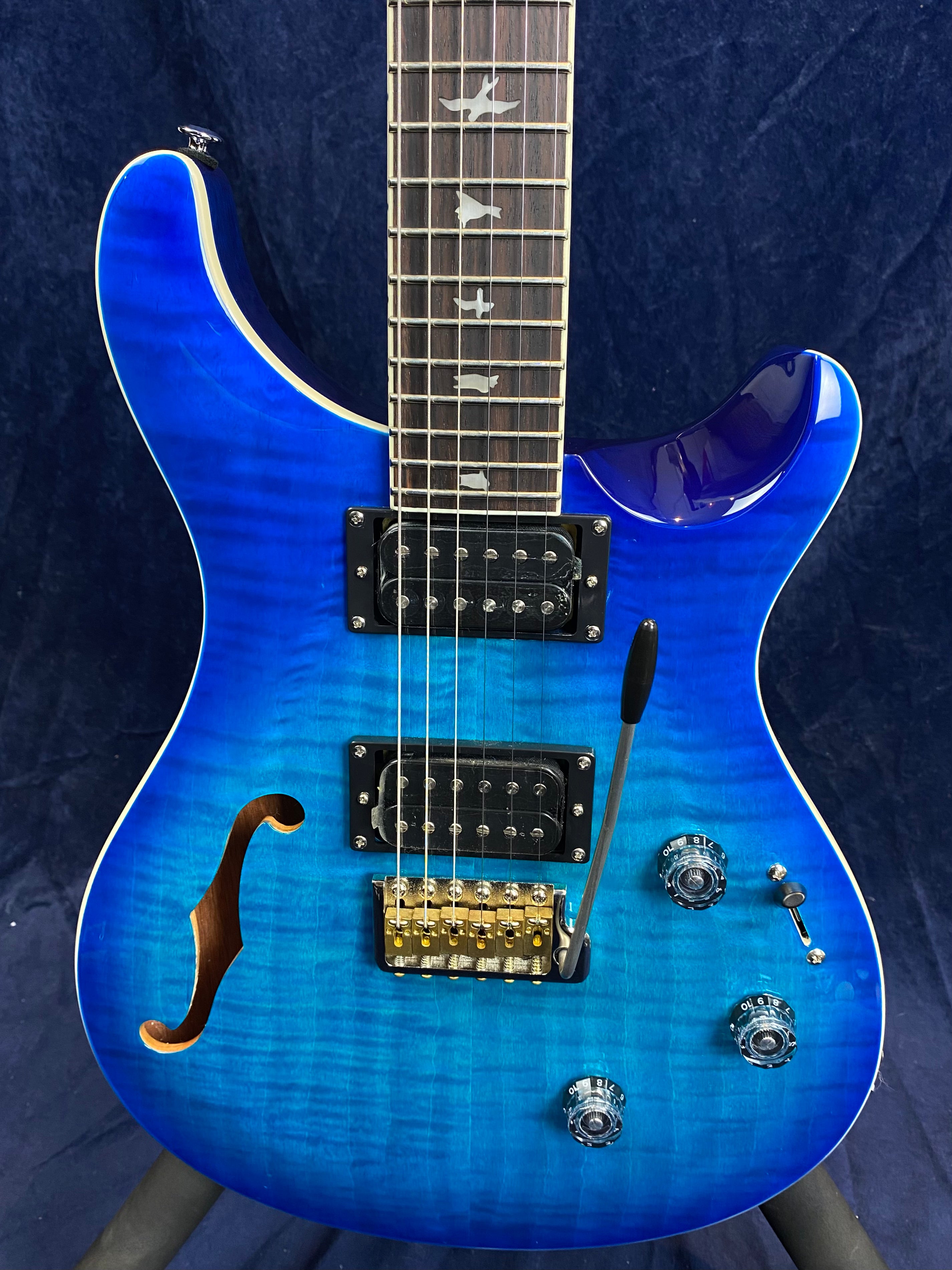 Bright blue electric guitar with a semi-hollow body, black dual humbucker pickups, transparent knobs, bird inlays on the fretboard, and tremolo bar.