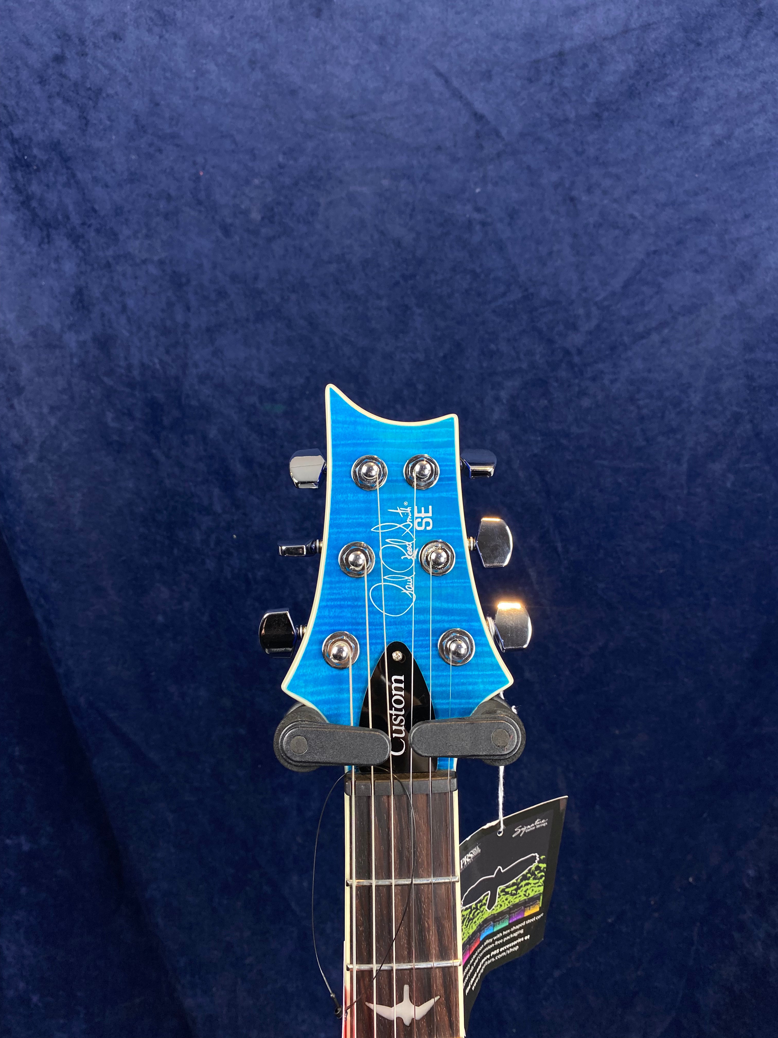 Blue electric guitar headstock with six chrome tuning pegs, signature, "Custom" truss rod cover, and a tag hanging from one tuning peg.