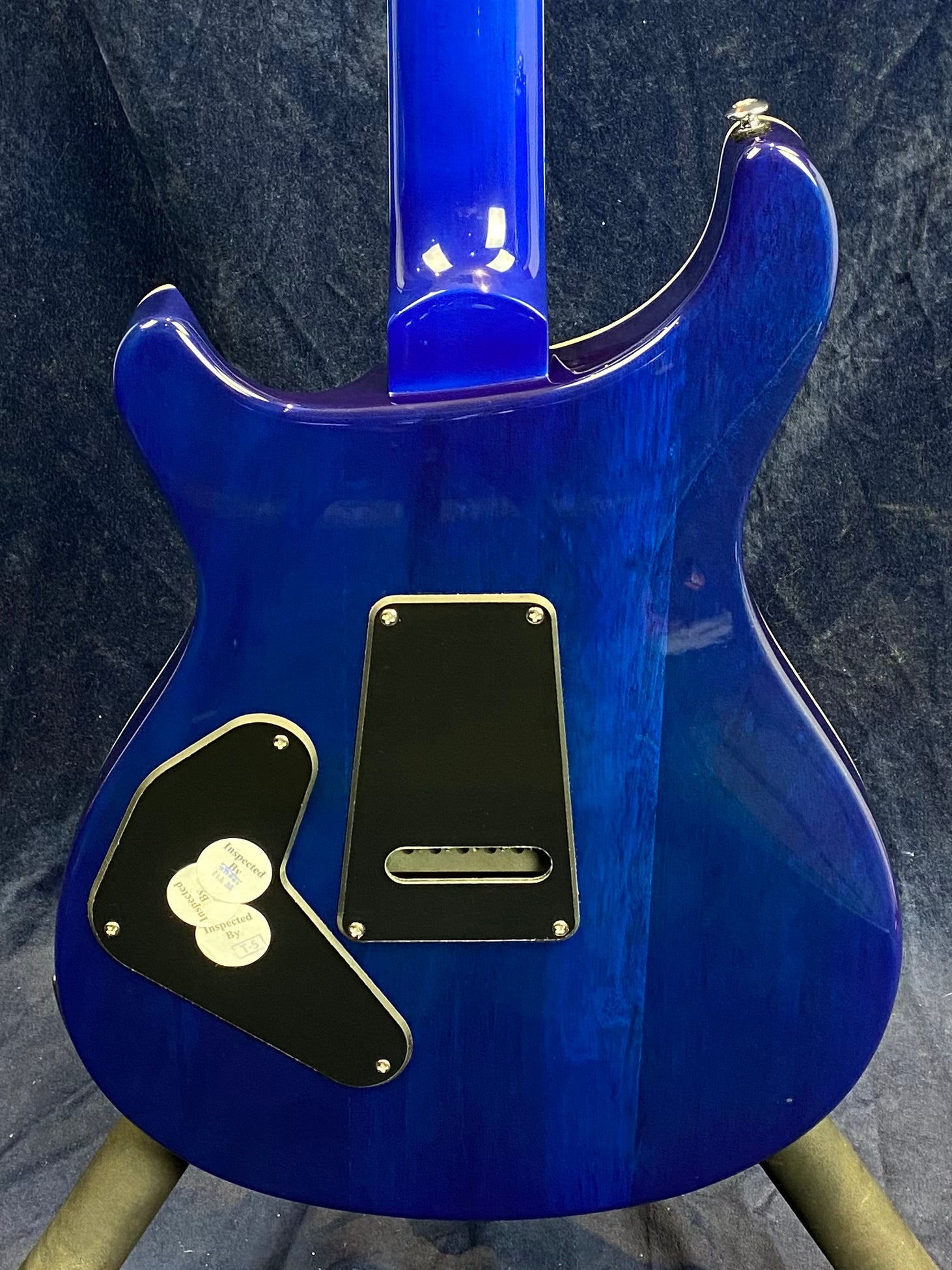 Rear view of a blue electric guitar body with two black backplates and three inspection stickers on the lower plate.
